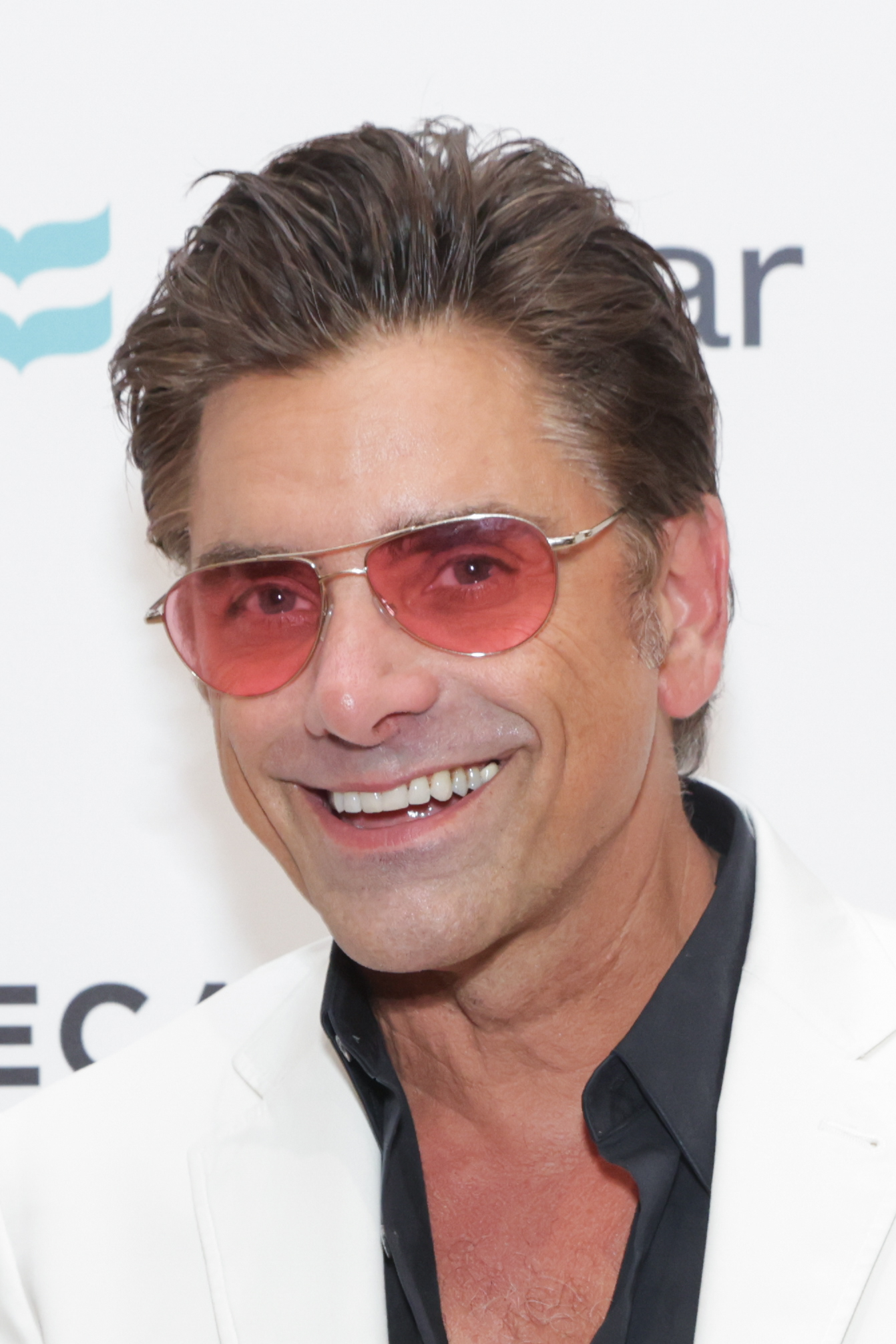John Stamos smiles, wearing a white blazer, black shirt, and red-tinted sunglasses at a public event