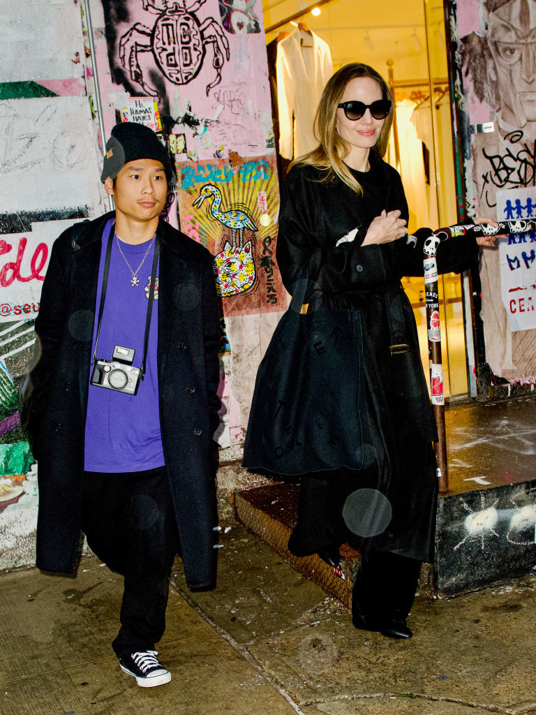 Angelina Jolie, in a black trench coat and sunglasses, stands next to a person wearing a purple shirt, black coat, and beanie. They are on a graffiti-covered street