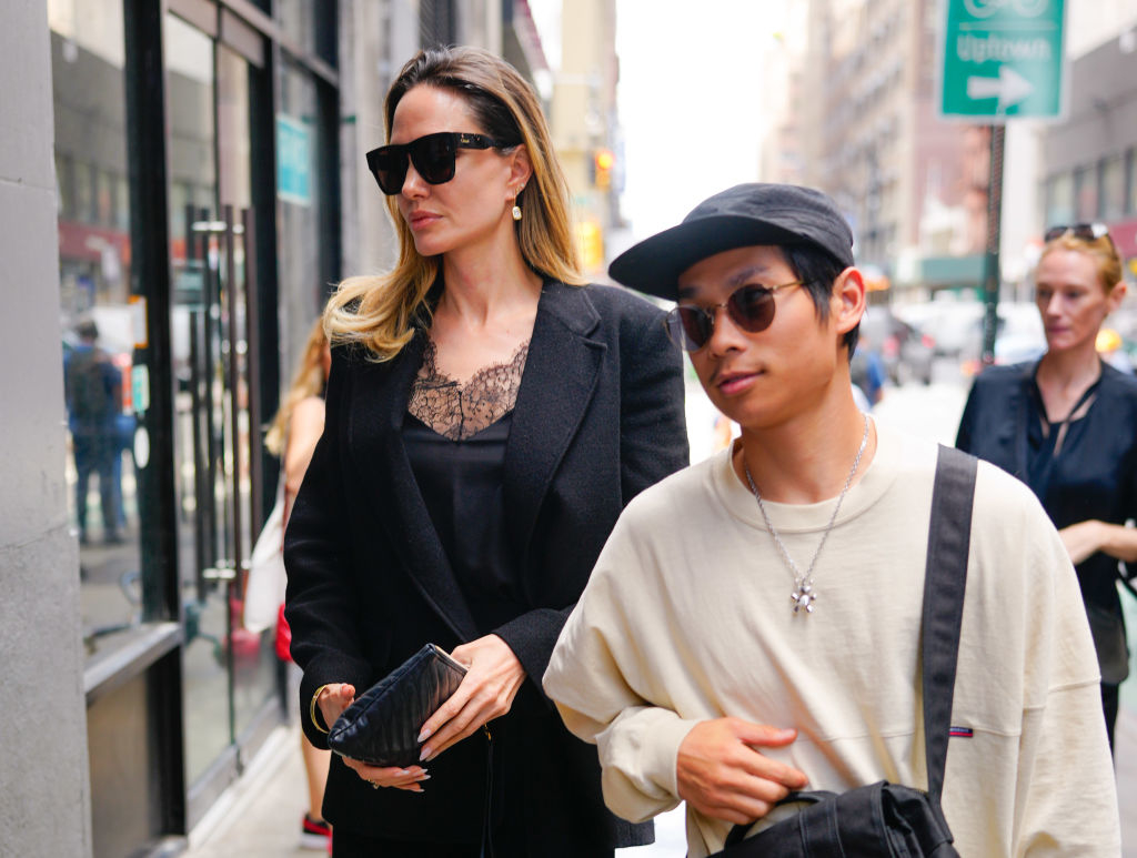 Angelina Jolie in a black lace-top dress and sunglasses walking with Prak behind her wearing a white sweater and baseball cap on a city street
