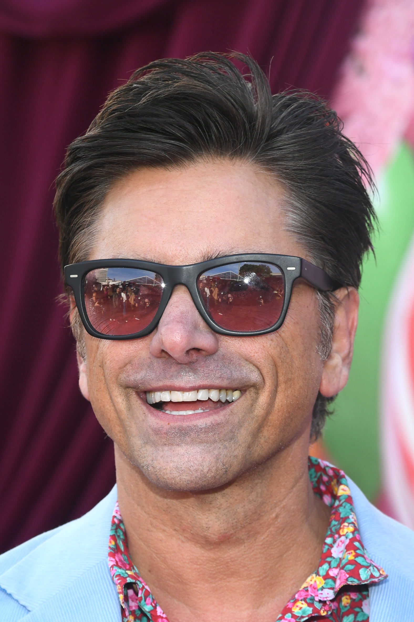 John Stamos, wearing sunglasses and a floral shirt, smiles at the camera