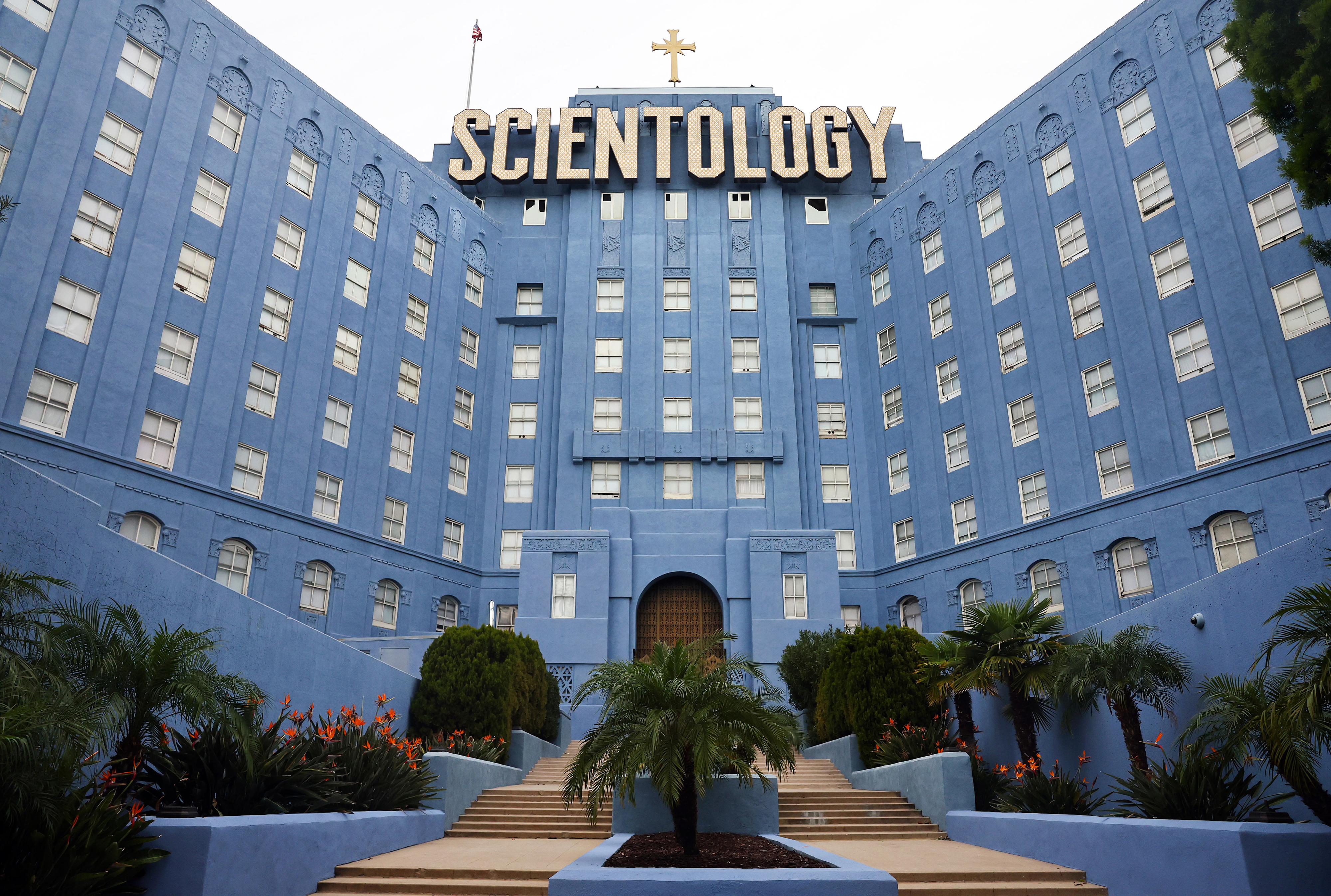 The image shows the front of the Church of Scientology building with expansive steps leading up to the entrance, surrounded by palm trees and greenery