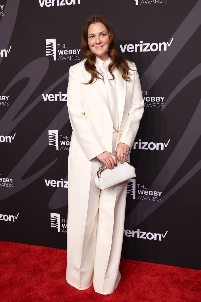 Drew Barrymore on the red carpet at The Webby Awards, wearing a white suit and holding a white clutch