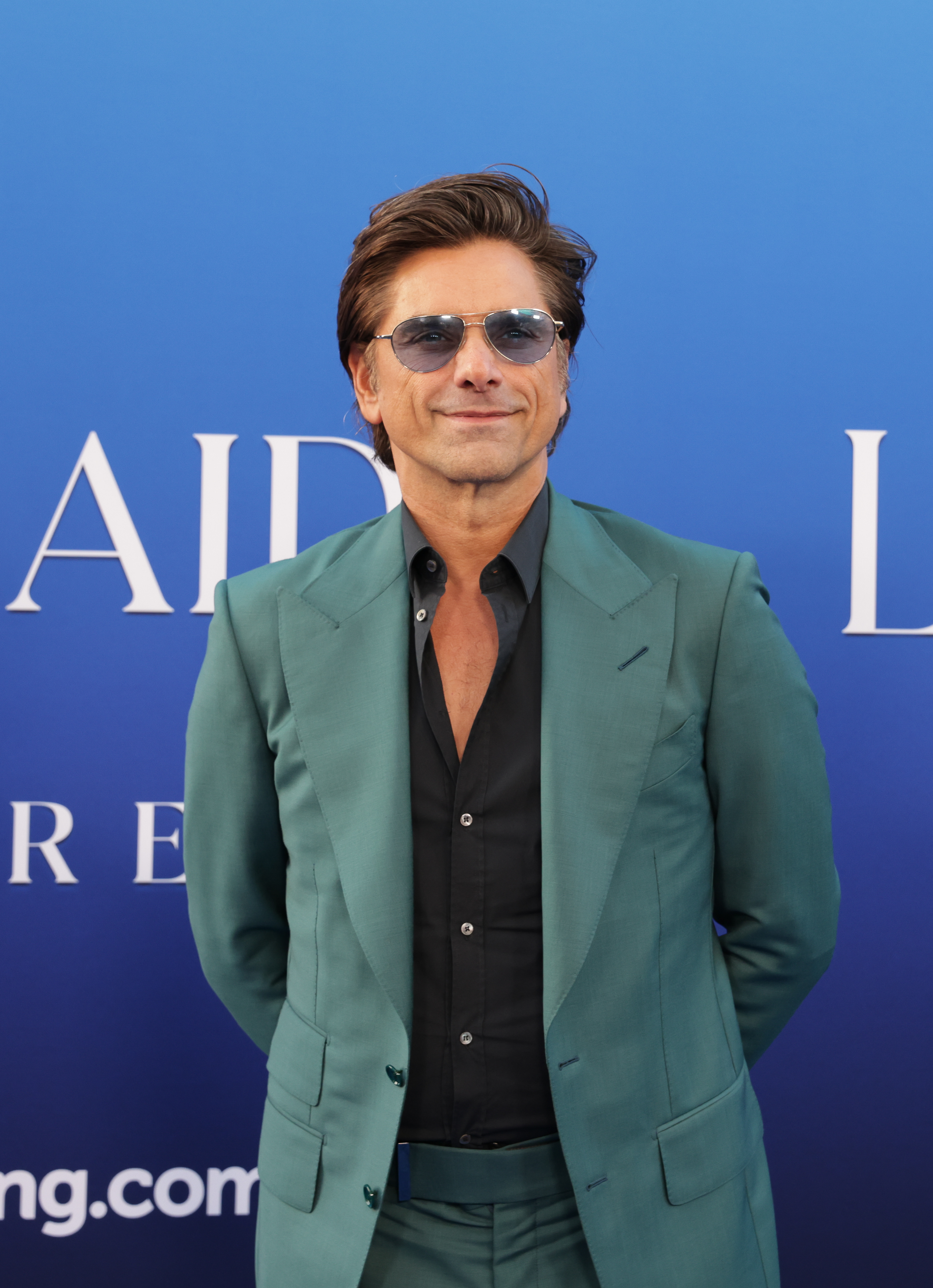 John Stamos poses in a teal suit and dark shirt at an event, standing in front of a blue background
