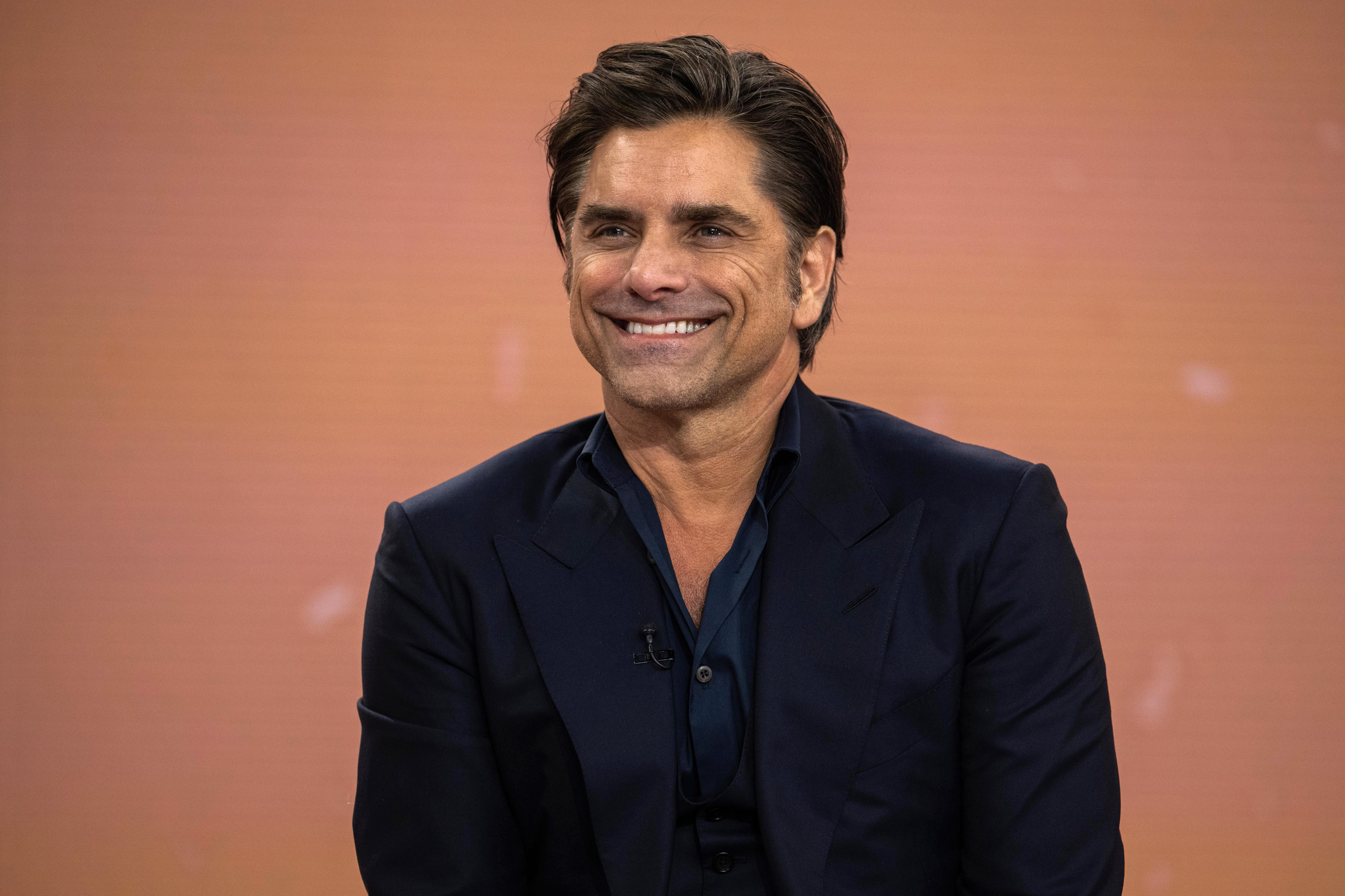 John Stamos smiles while seated in a dark suit with an open-collar shirt. The background is plain