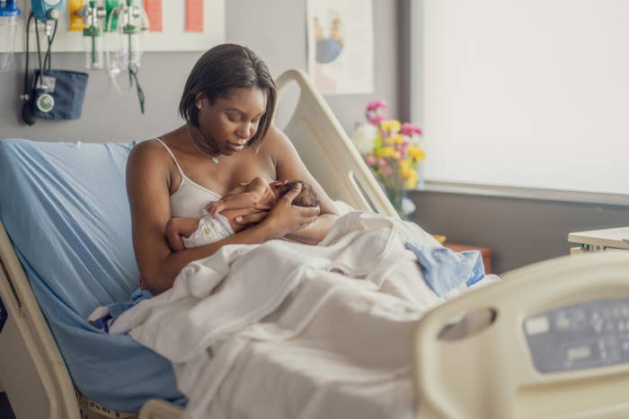 A mother gently holds and breastfeeds her newborn baby while sitting on a hospital bed, surrounded by medical equipment and colorful flowers