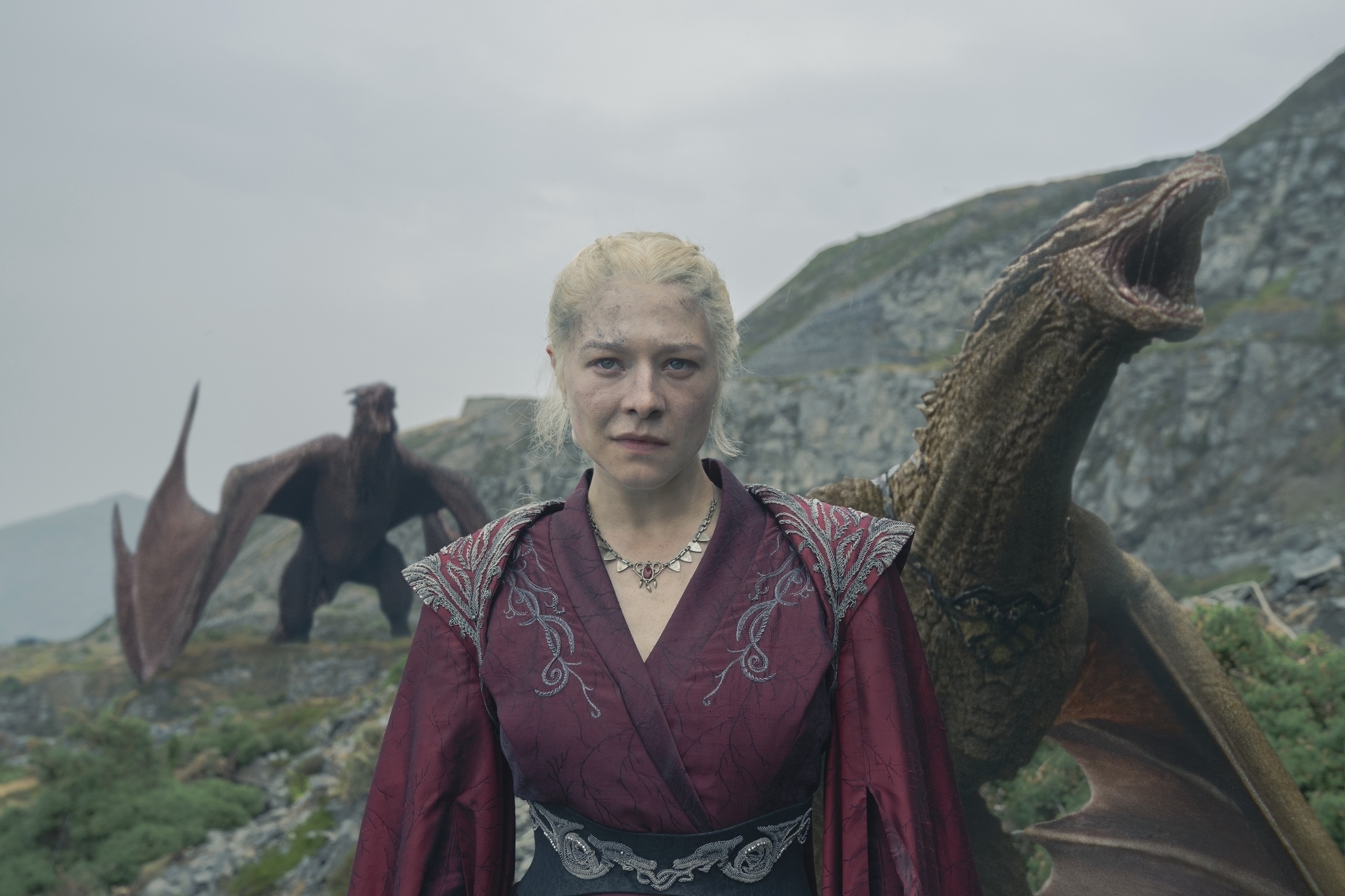 Emma D'Arcy, in elaborate medieval-style attire, stands between two dragons against a rocky landscape