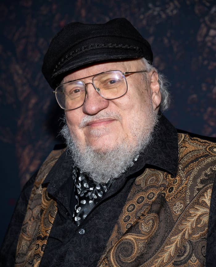 George R. R. Martin is wearing a paisley vest over a dark shirt, a patterned tie, and a black cap. He has a white beard and large glasses
