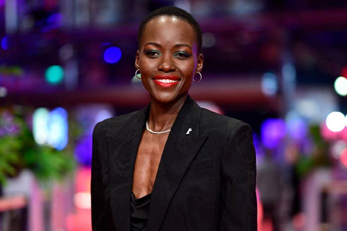 Lupita Nyong'o on a red carpet, smiling, wearing a black blazer over a black top, accessorized with a necklace and hoop earrings
