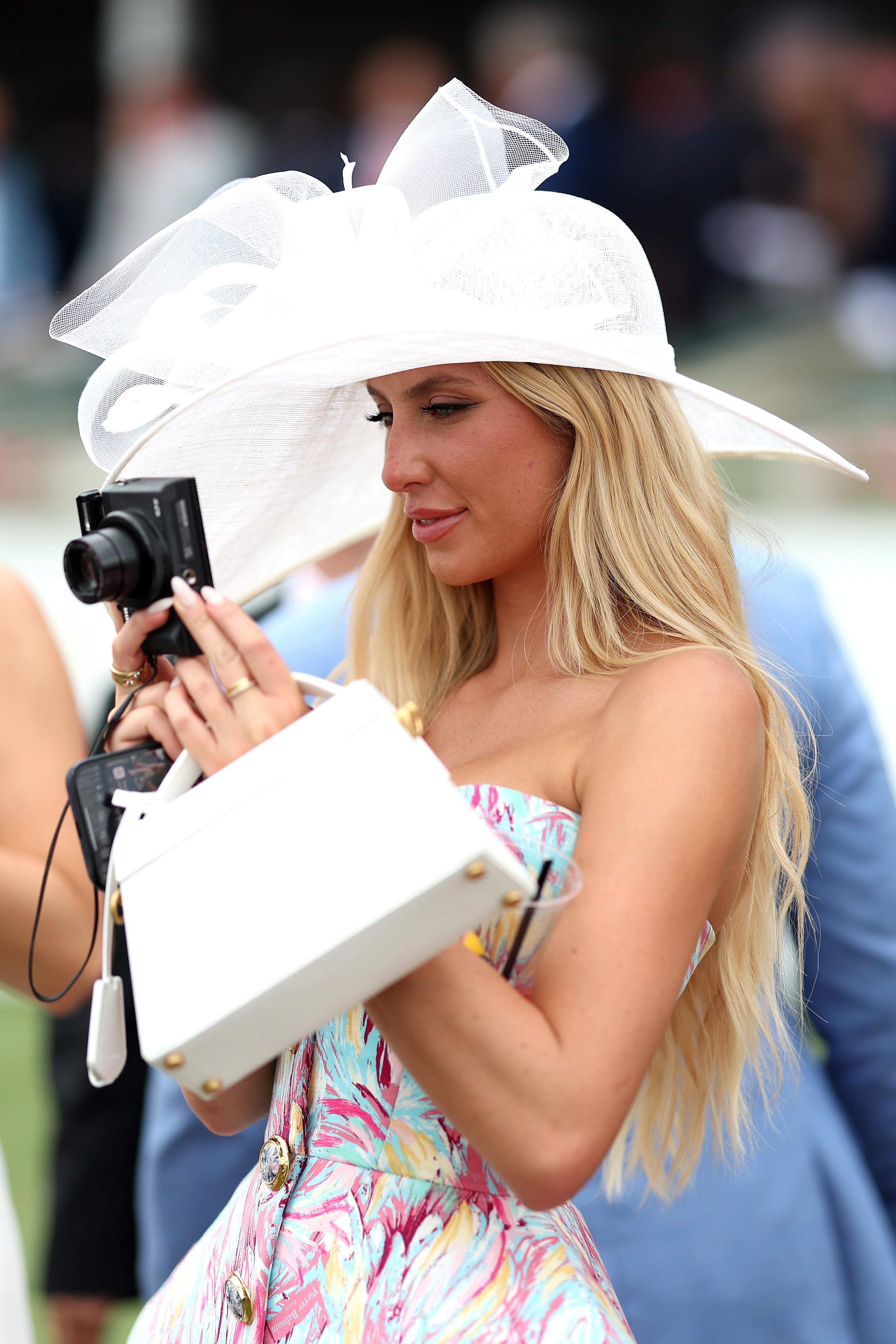 A woman dressed in a stylish, floral print dress and large hat holds a camera, capturing photos at an outdoor event