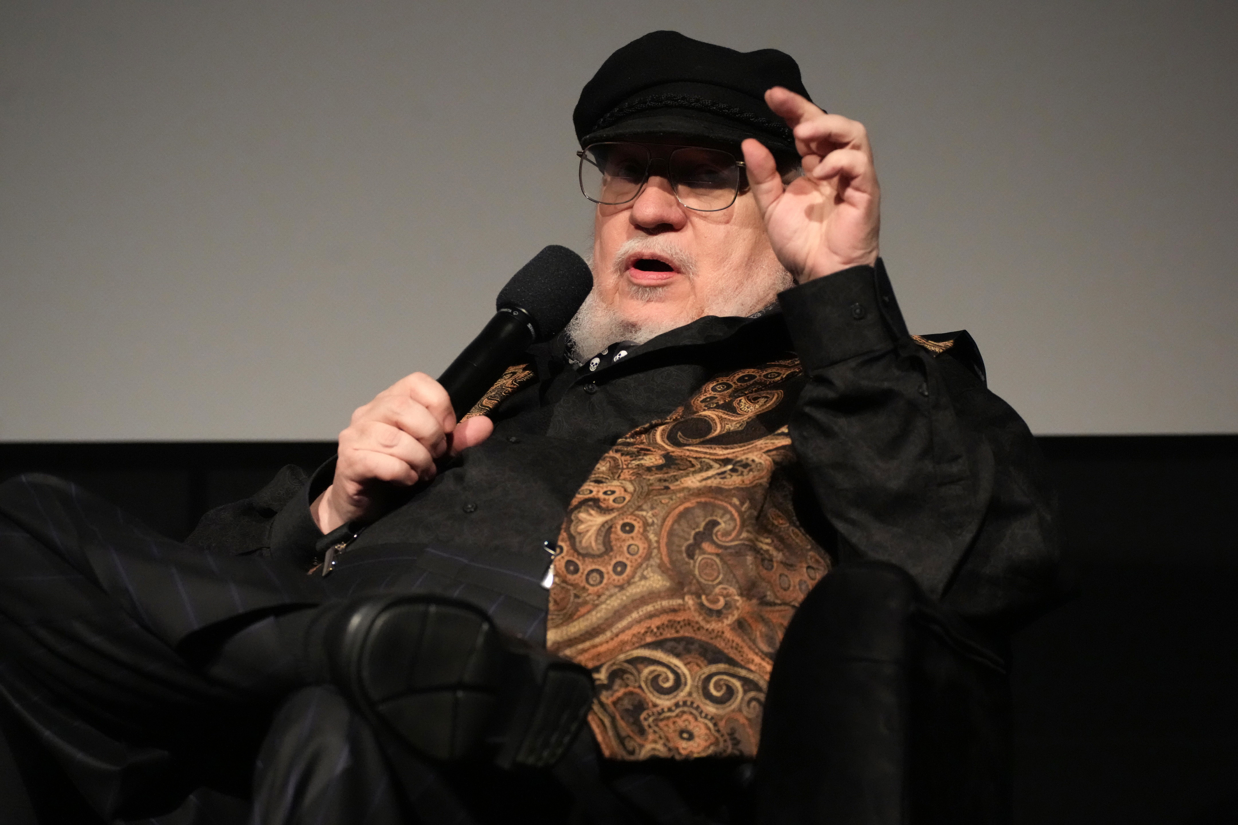 George R. R. Martin, author, wearing a patterned vest and black cap, speaks into a microphone while gesturing with his hand