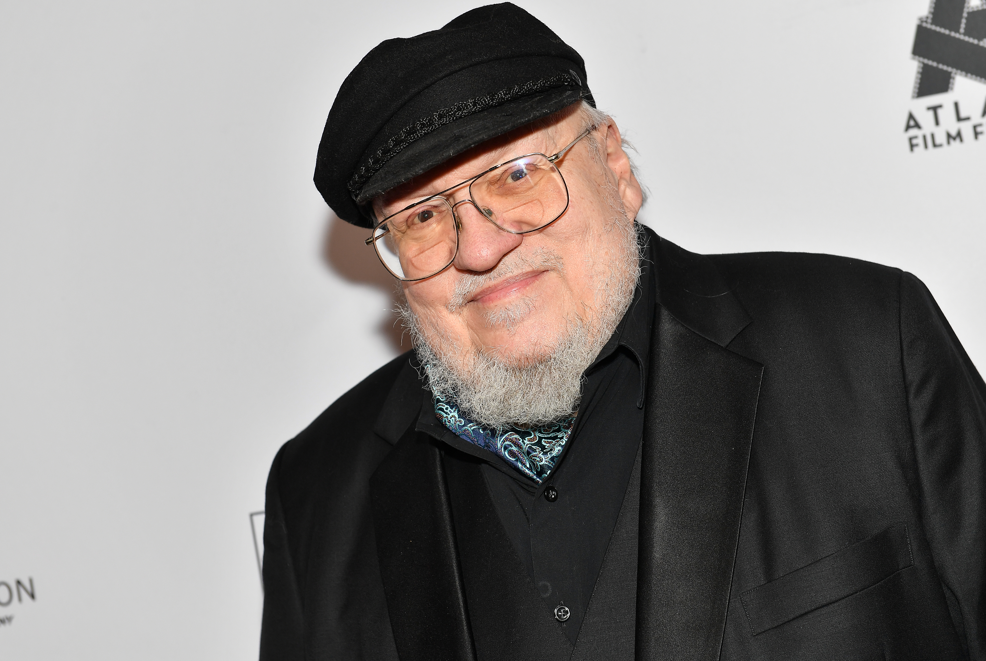George R.R. Martin smiles while wearing a black suit, a black cap, and a bandana, attending an event