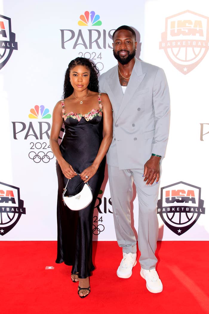 Gabrielle Union, in a black satin dress with floral accents, and Dwyane Wade, in a light gray suit with white sneakers, on a red carpet at a Paris 2024 event