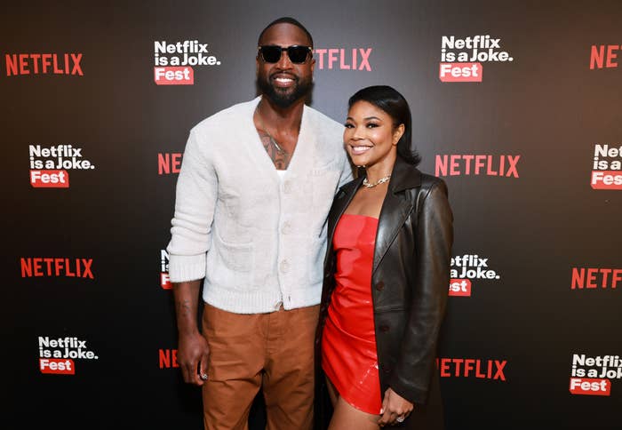 Dwyane Wade in a casual cardigan and sunglasses, and Gabrielle Union in a chic red dress with a brown coat, pose together at a Netflix event backdrop
