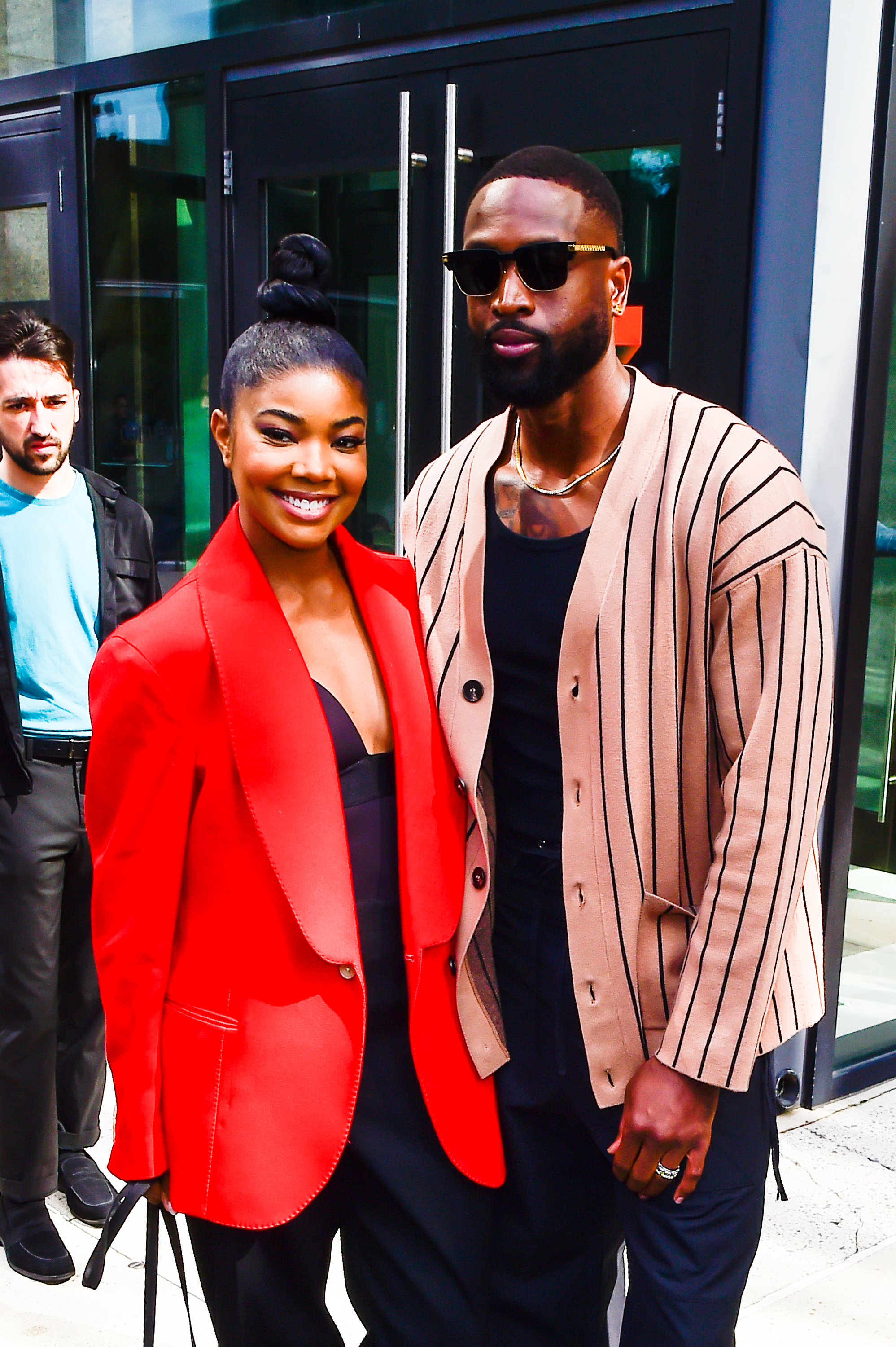 Gabrielle Union in a chic red blazer and Dwyane Wade in a stylish striped cardigan and sunglasses, smiling together outside a building