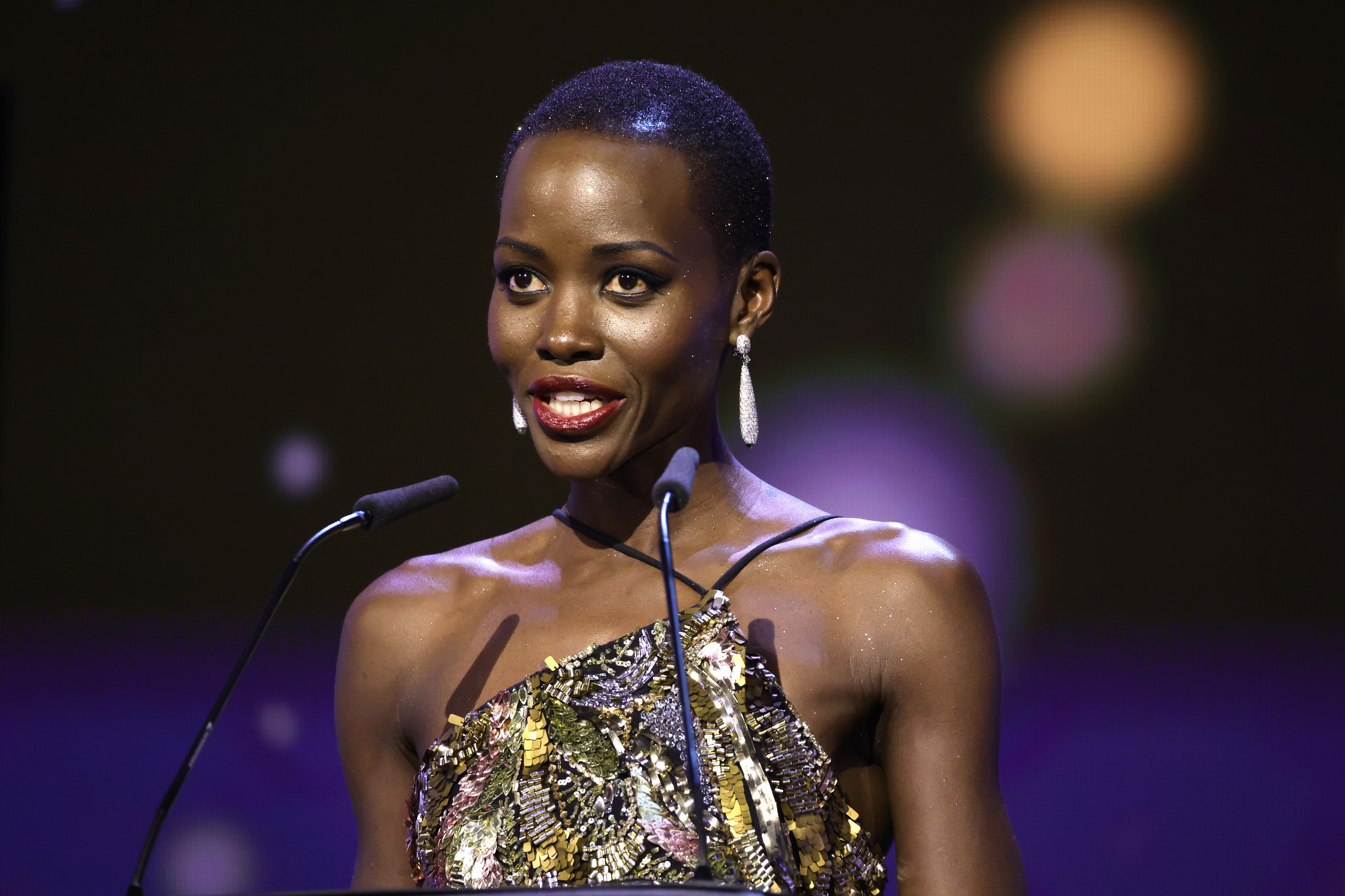 Lupita Nyong'o speaks at an event, wearing an elegant one-shoulder dress with a sparkly, textured pattern and long earrings