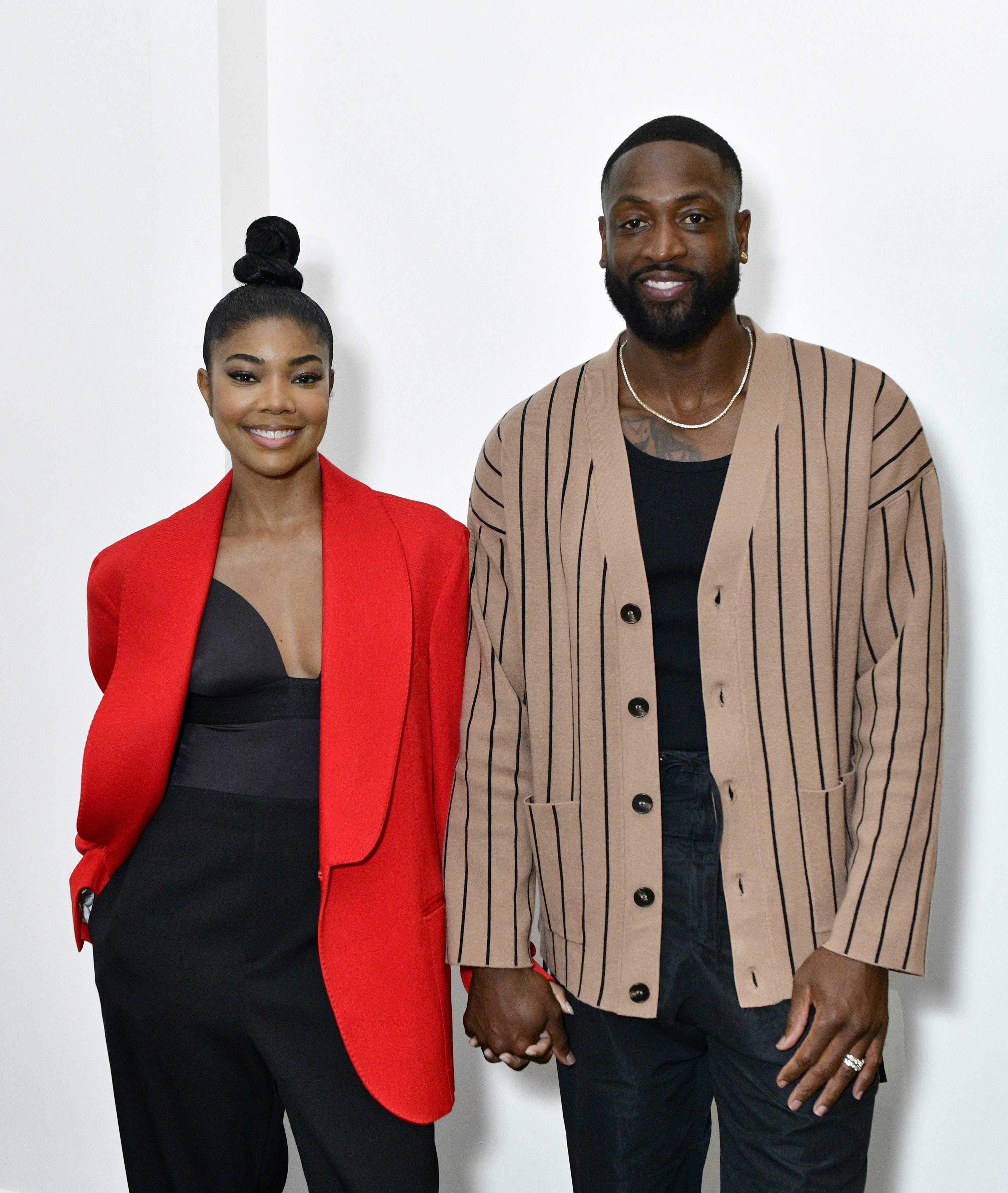 Gabrielle Union and Dwyane Wade pose together. Gabrielle wears a red blazer and black top, while Dwyane wears a beige cardigan with vertical stripes