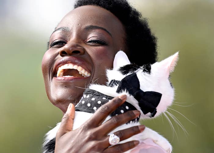 Lupita Nyong'o smiles broadly while holding a black-and-white cat wearing a polka-dot bow tie