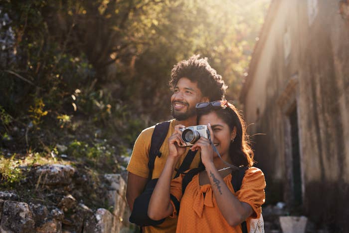 Man with a beard and woman with a tattoo and camera, both wearing backpacks, smile while exploring a sunlit outdoor area with foliage and an old building