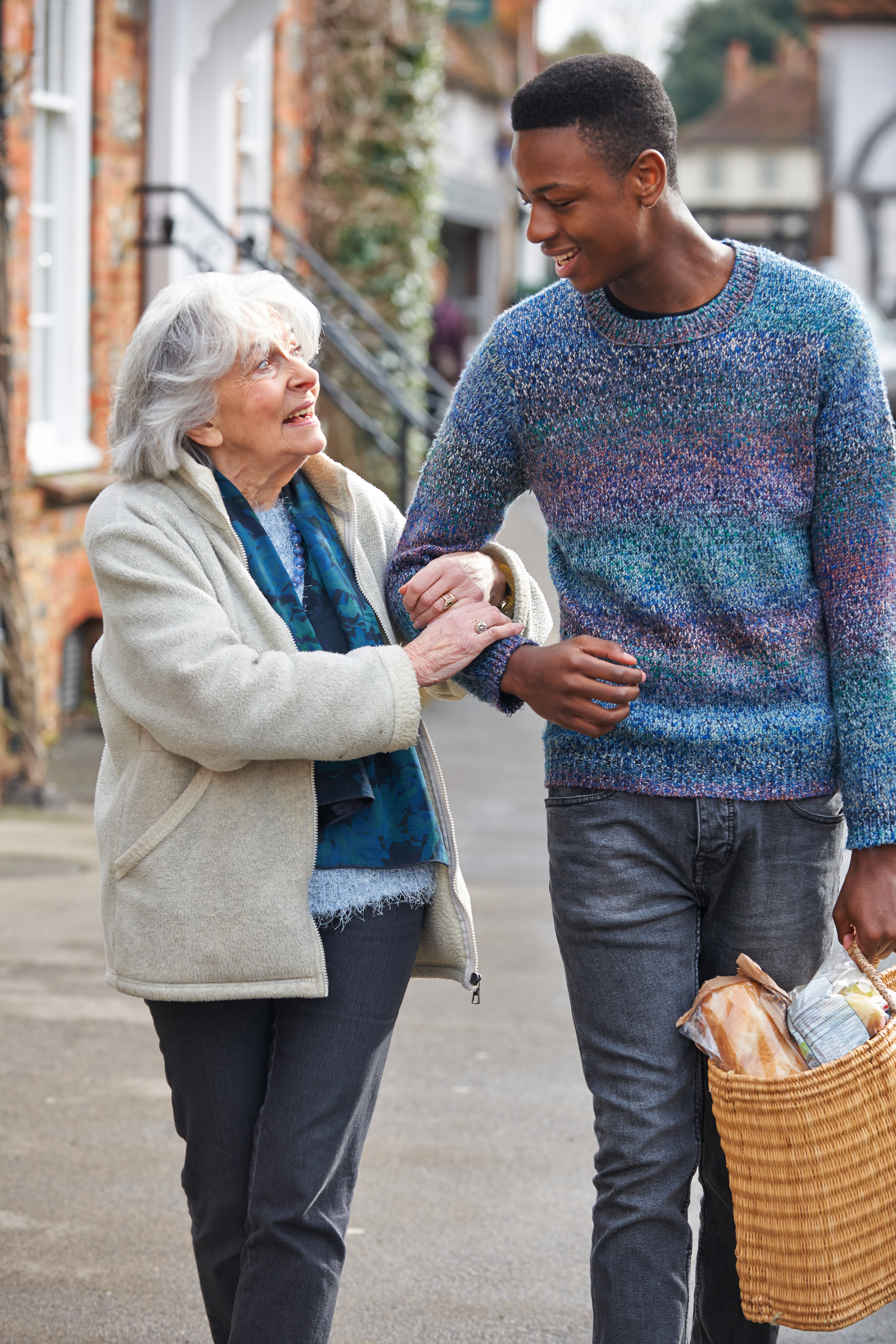 An elderly woman and a young man smile as they walk arm-in-arm on a sidewalk while carrying grocery bags