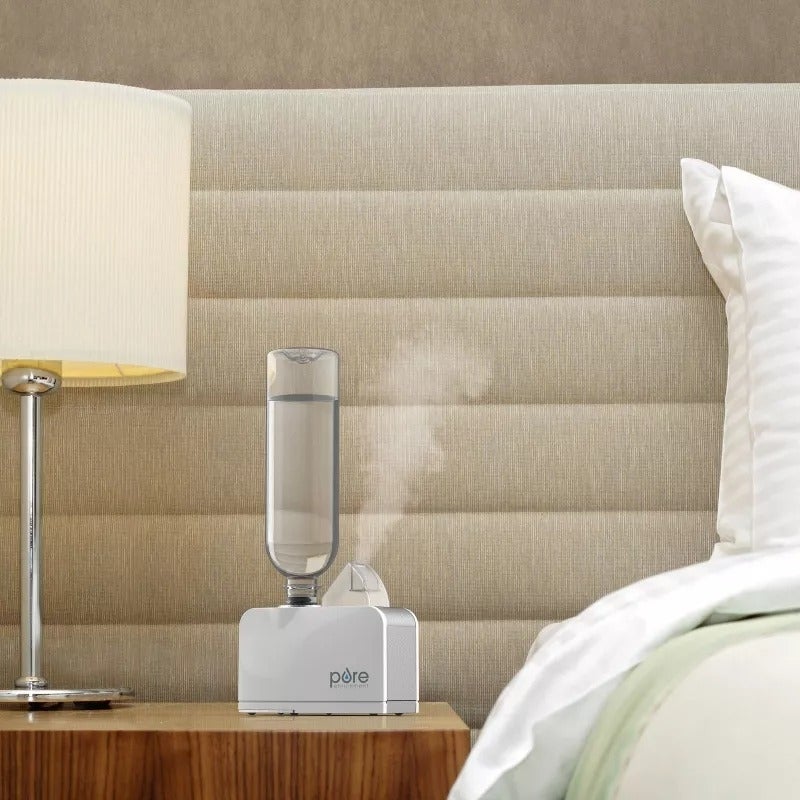 A modern   chamber  country   featuring a achromatic  Pure humidifier connected  a nightstand adjacent  to a lit array  lamp. A beige upholstered headboard and portion  of a furniture  are visible
