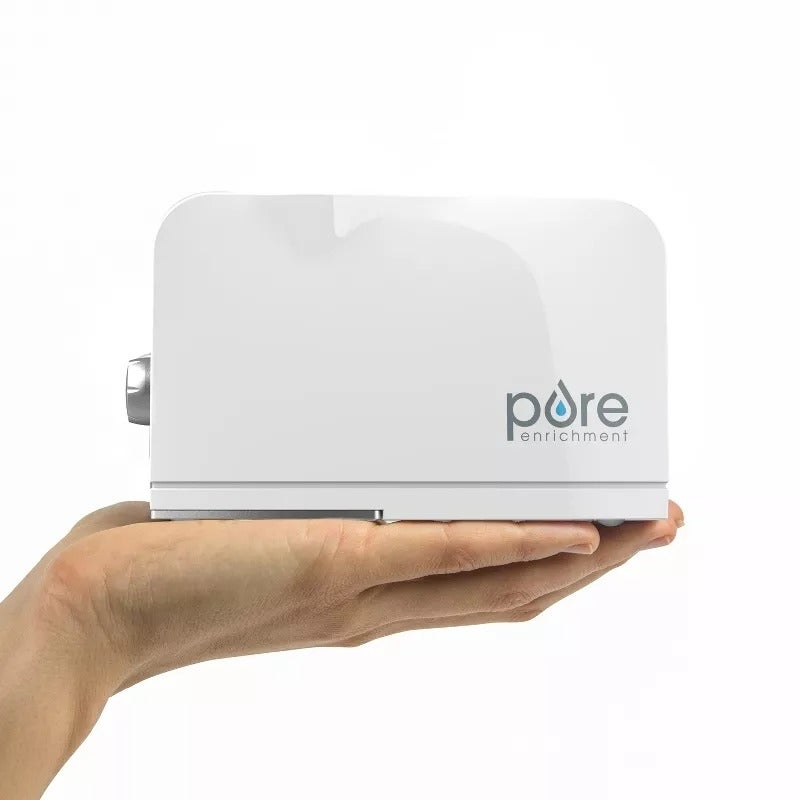 A manus  holds a compact achromatic  Pure Enrichment humidifier with the marque  logo visible. The instrumentality  is tiny  and sleek, suitable for location  use