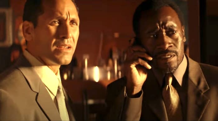 Two unnamed individuals are depicted seriously conversing; one is on the phone. Both are dressed in professional suits, in likely indoor or office setting