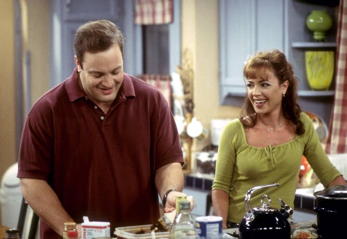 Kevin James and Leah Remini are in a kitchen scene. Kevin is smiling while Leah, wearing a casual off-shoulder top, looks at him