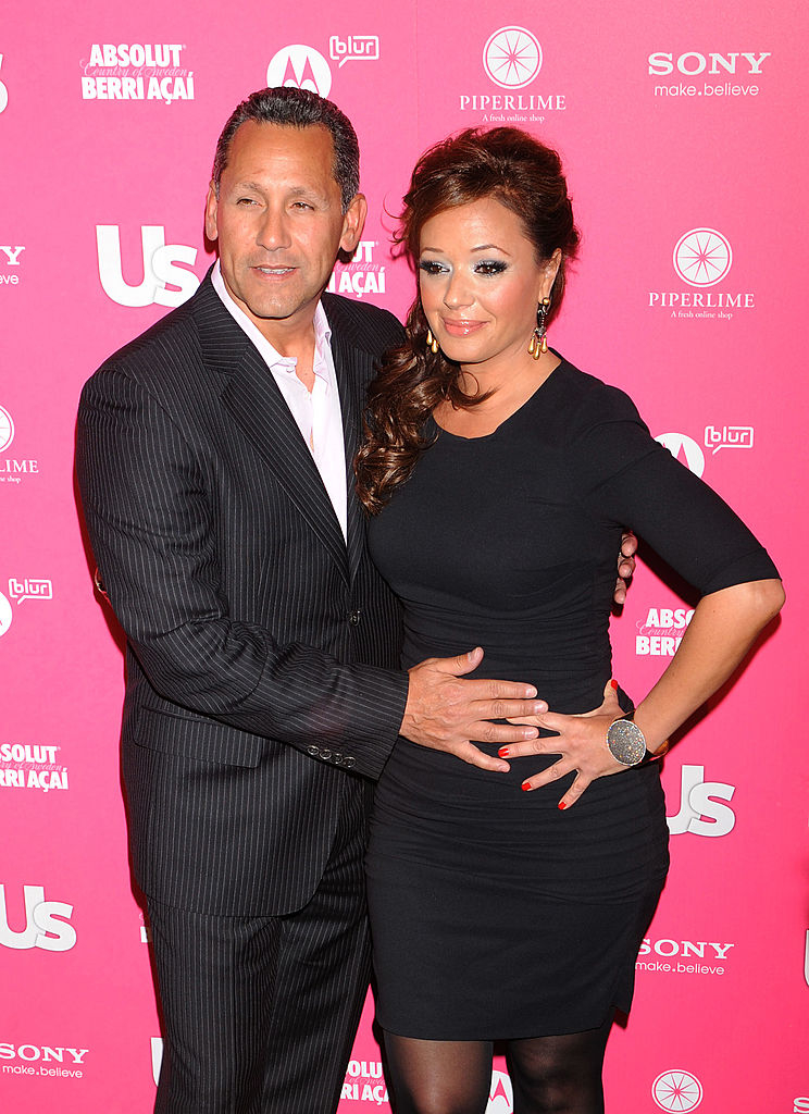 A man in a dark suit and Leah Remini in a fitted black dress smile on a red carpet at a Us Weekly event