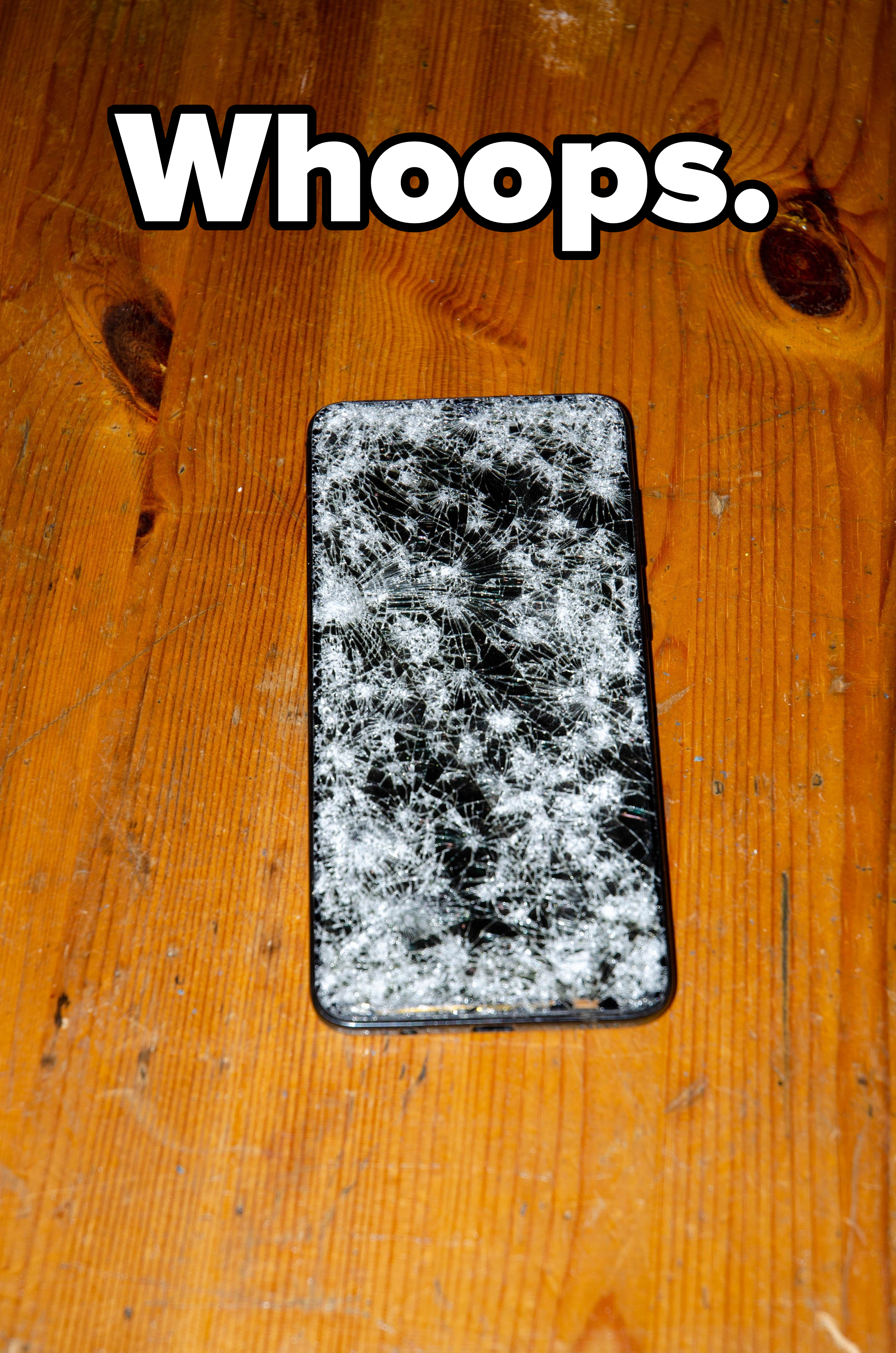 A shattered smartphone screen lies face-up on a wooden surface