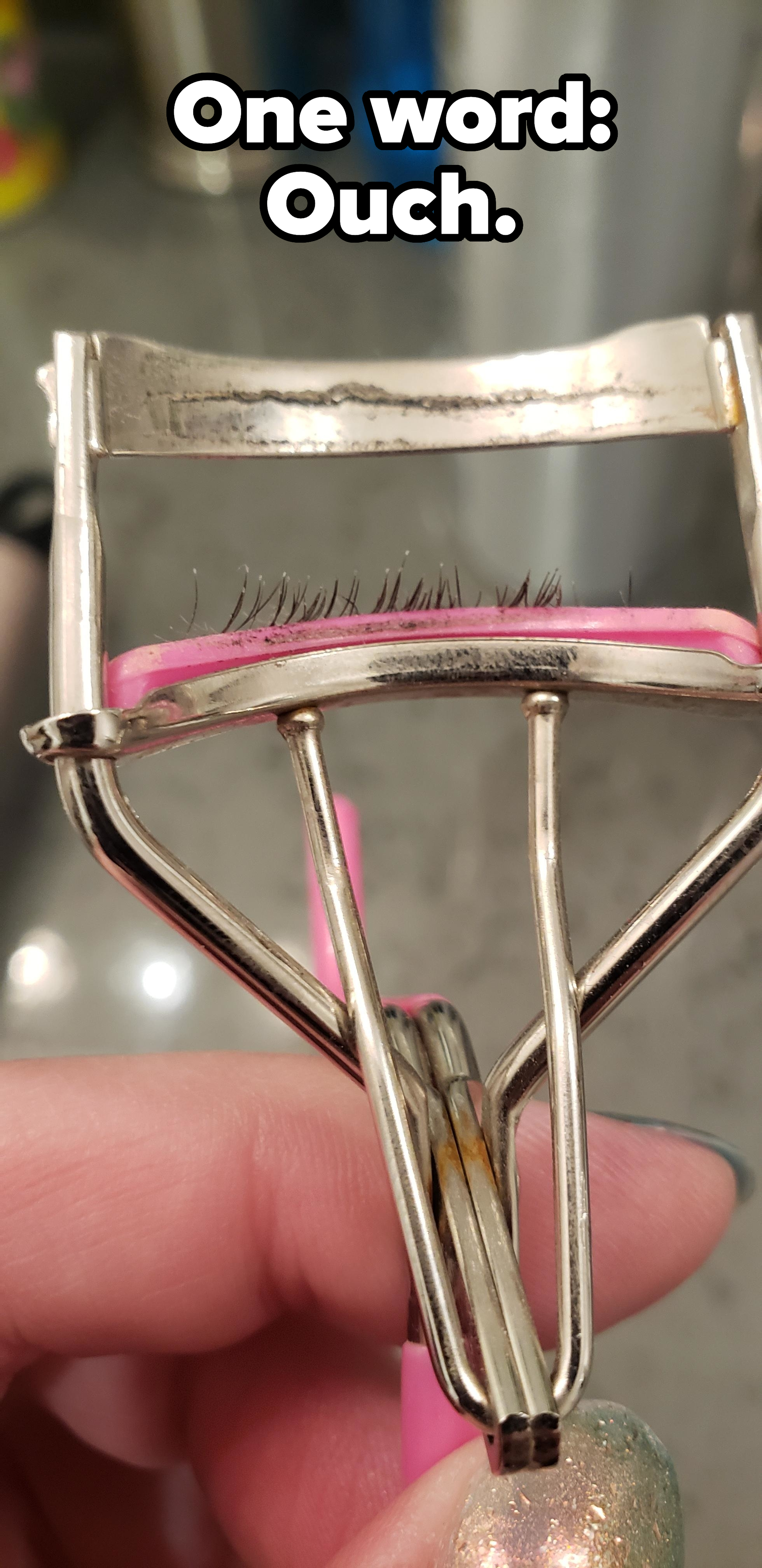 A close-up of a metallic eyelash curler held by a person's hand, showing eyelashes caught in the curler