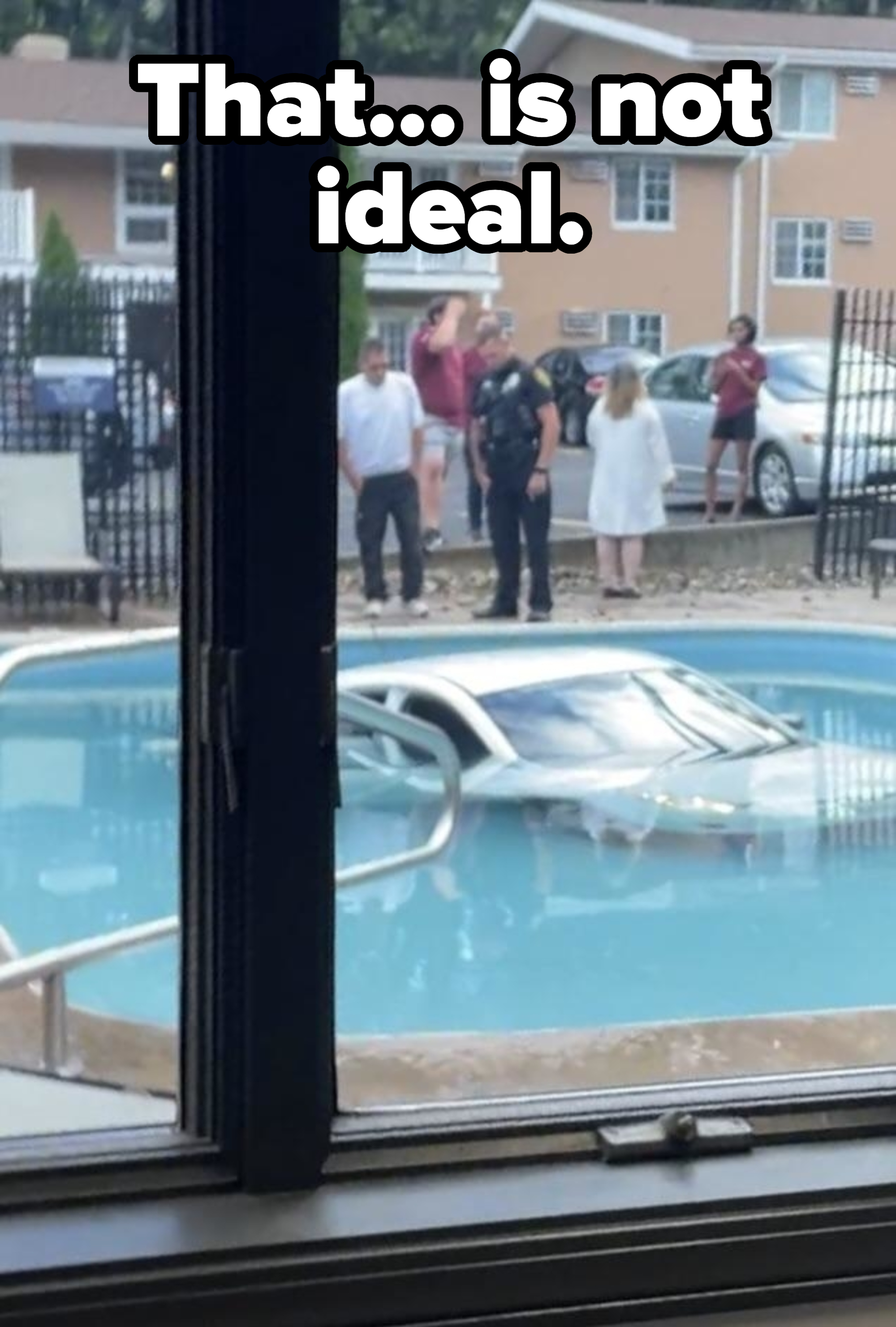 A car is partially submerged in a pool. Nearby, a police officer and several people stand by, observing the scene. Apartments and parked cars are in the background