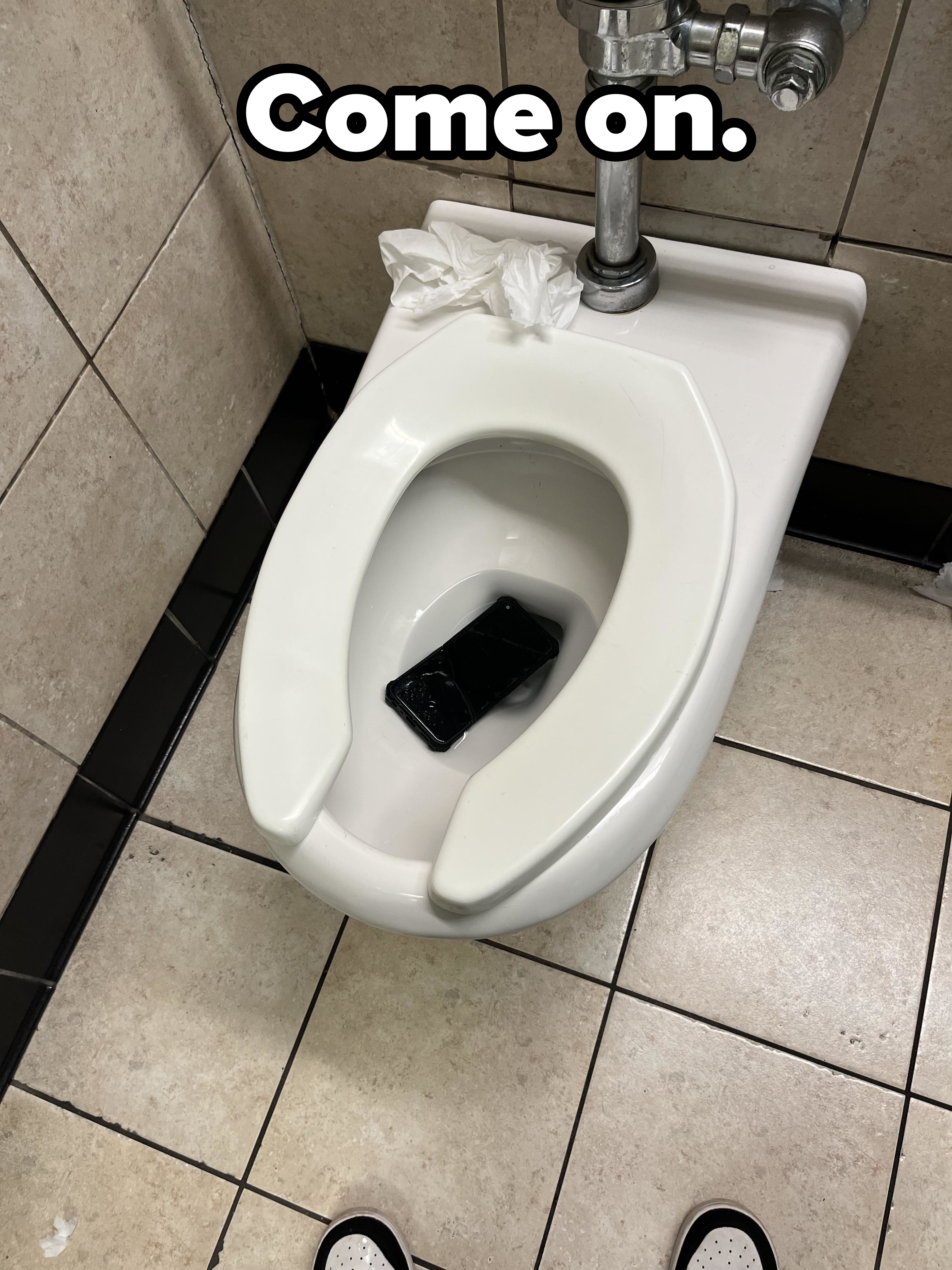 A smartphone is submerged in the water of a toilet bowl with a few pieces of tissue resting on the rear part of the seat