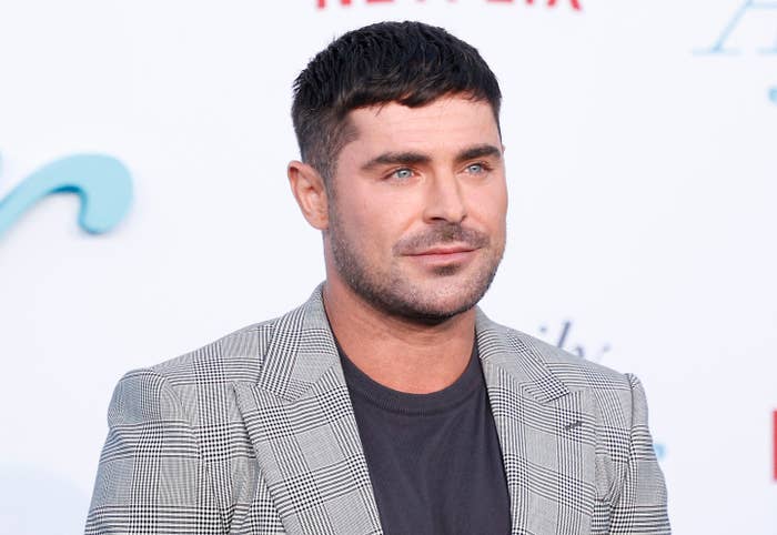 Zac Efron poses at a media event in a checkered suit jacket and black shirt, with a neutral facial expression