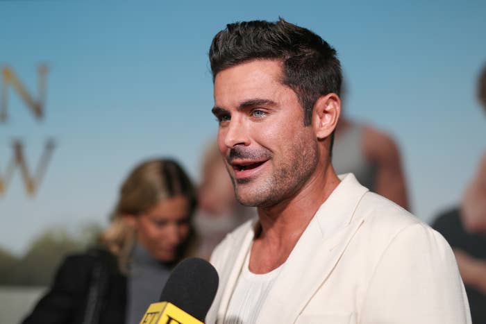 Zac Efron in a light-colored suit talks to reporters at a press event, holding a microphone. Background shows blurred people