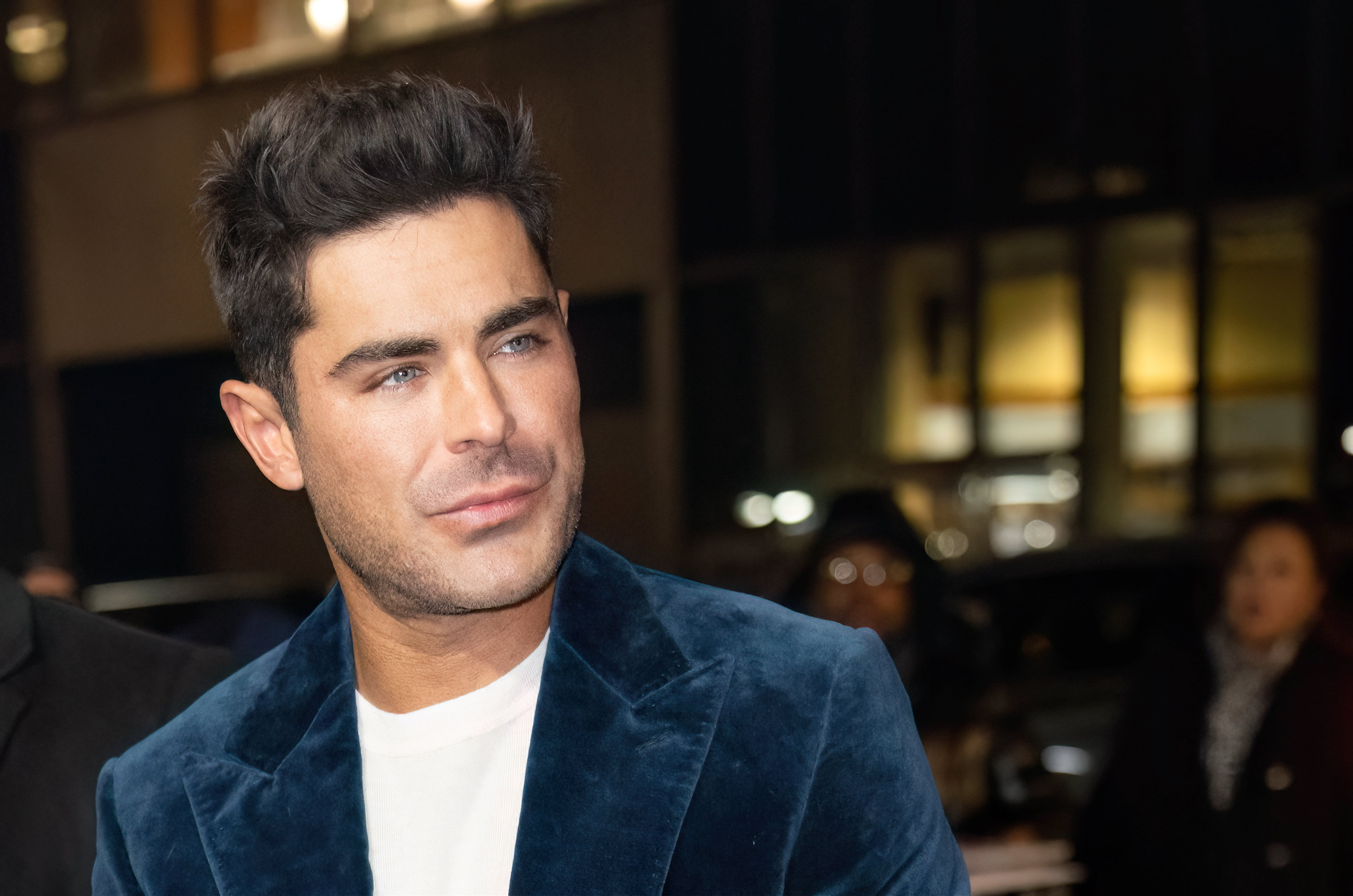 Zac Efron attends an event wearing a white shirt and a velvet blazer