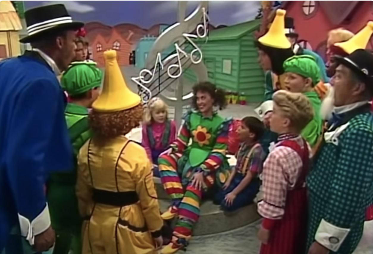 Group of people in colorful costumes, including clowns and characters with tall hats, gathered around a seated individual with a flower on their shirt