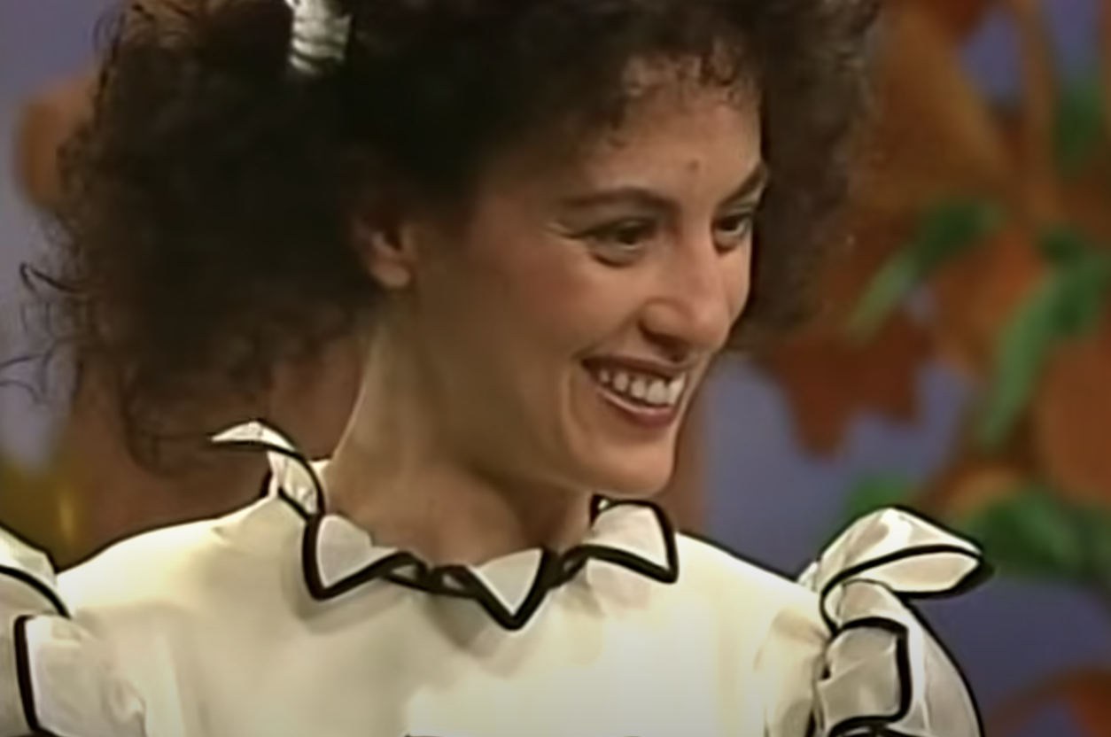 A woman with curly hair smiles while wearing a white dress with black trim along the collar and sleeves