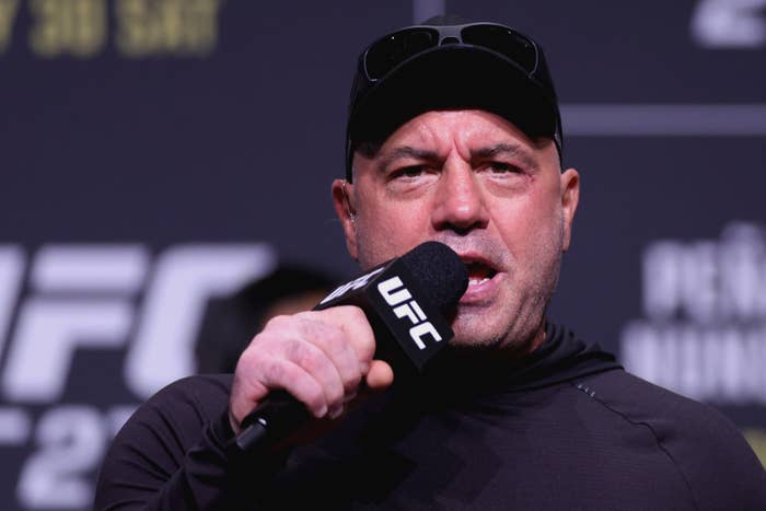 Joe Rogan speaking into a UFC-branded microphone at an event, wearing a cap and casual attire