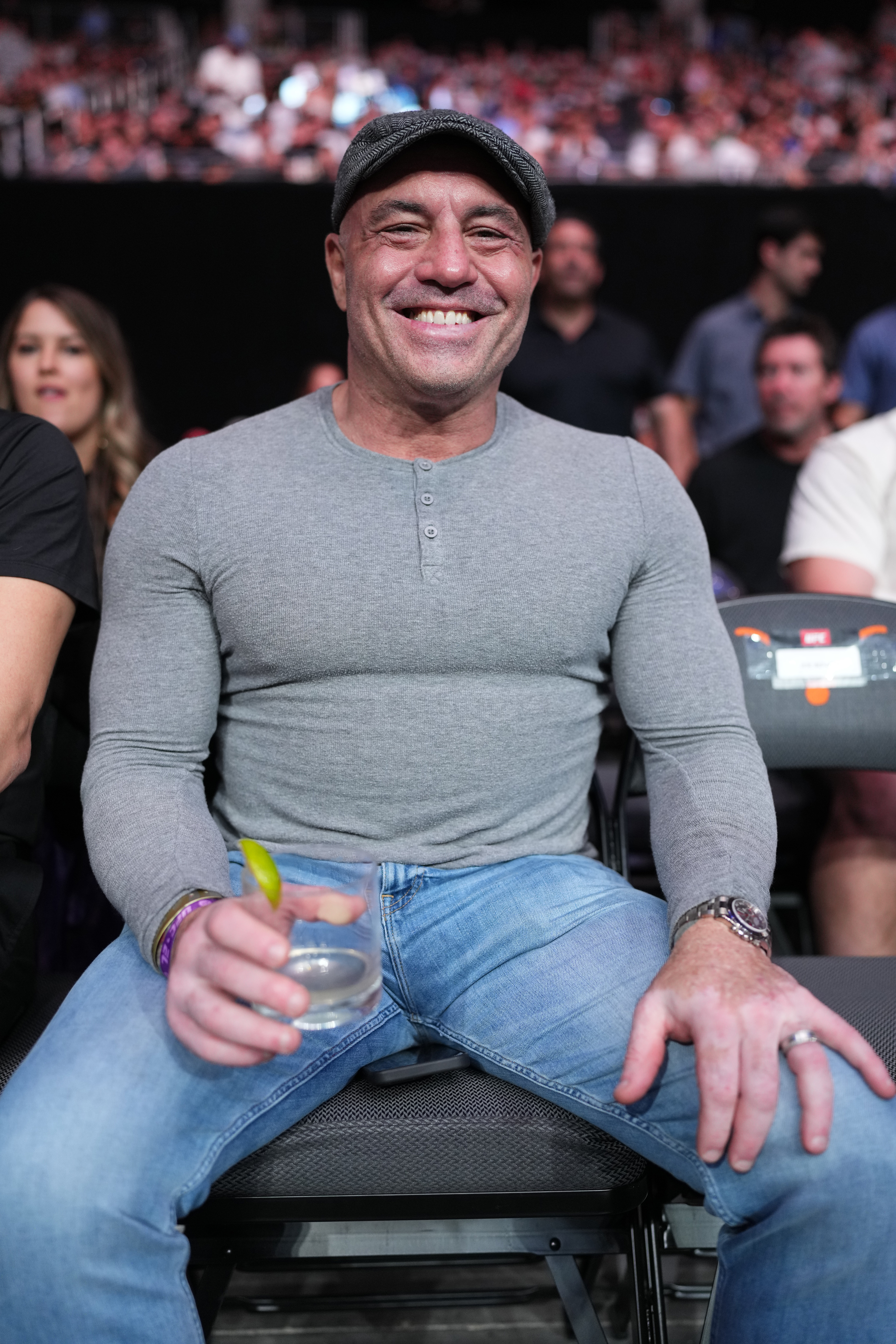 Joe Rogan is sitting in an audience, smiling, wearing a grey long-sleeve shirt, blue jeans, and a cap, holding a beverage with a lime