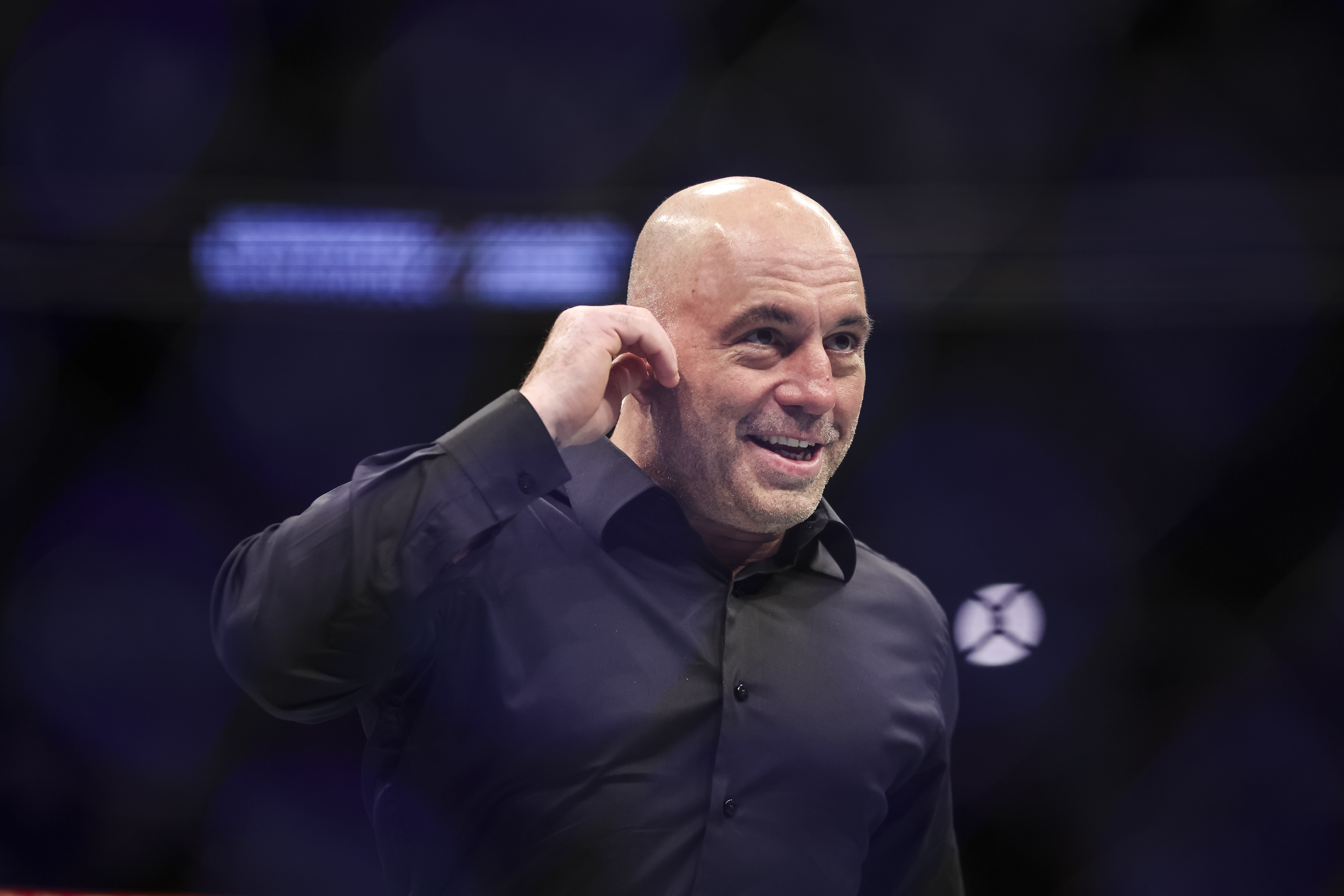 Joe Rogan, wearing a dark shirt, is seen smiling and touching his ear on stage