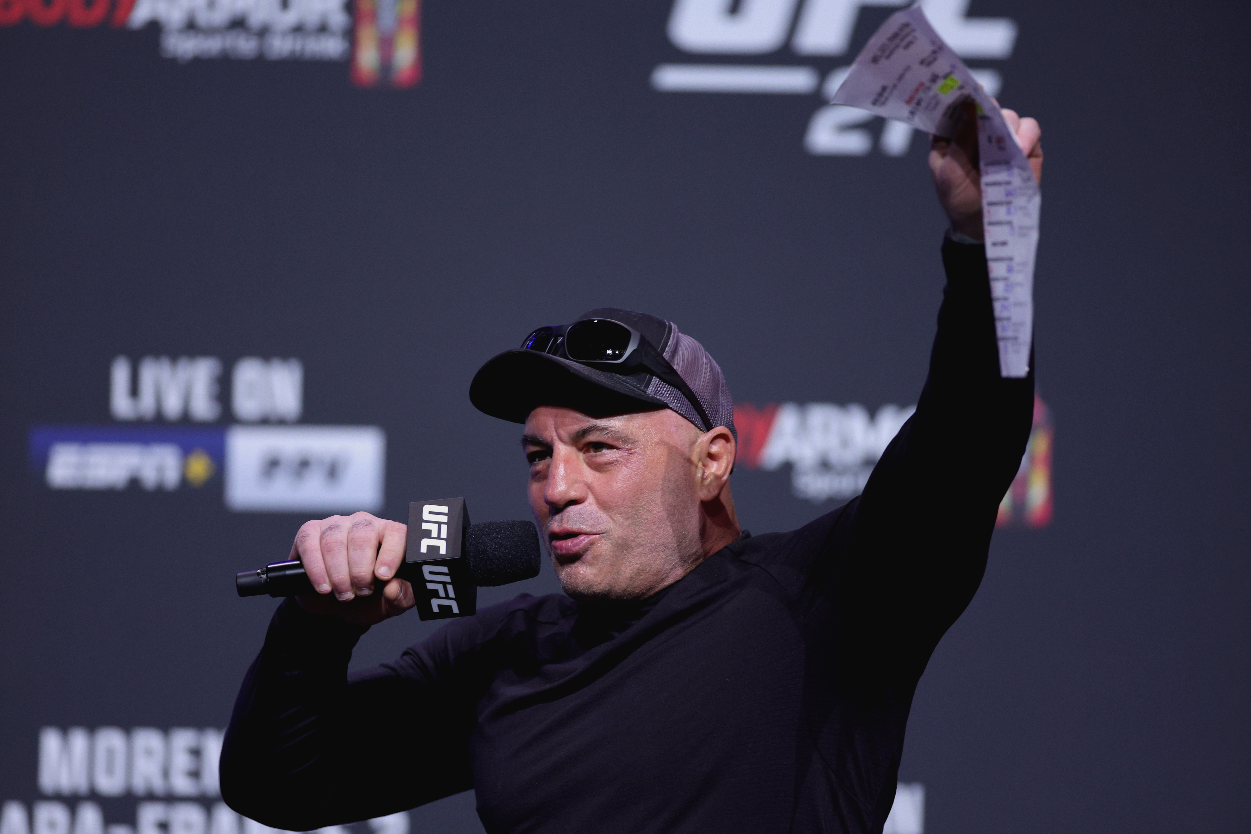 Joe Rogan on stage holding a microphone and a sheet of paper, speaking at a UFC 272 event