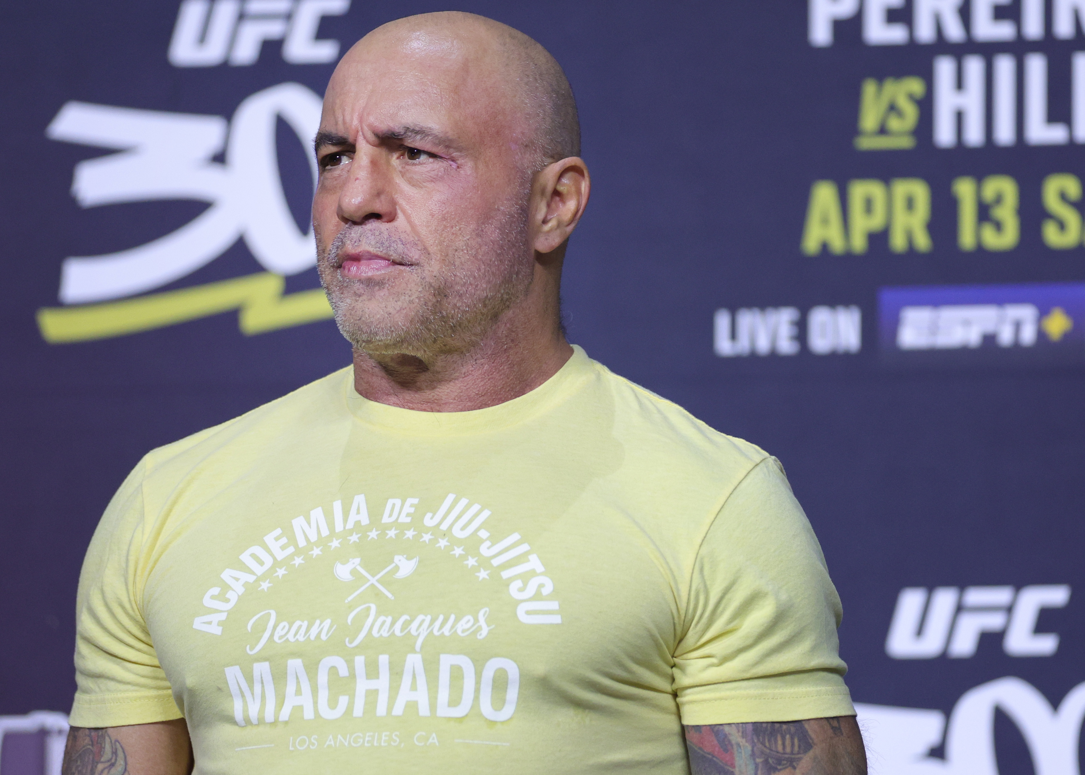 Joe Rogan at a UFC event wearing a yellow t-shirt with "Académie Jiu-Jitsu, Jean Jacques Machado, Los Angeles, CA" printed on it