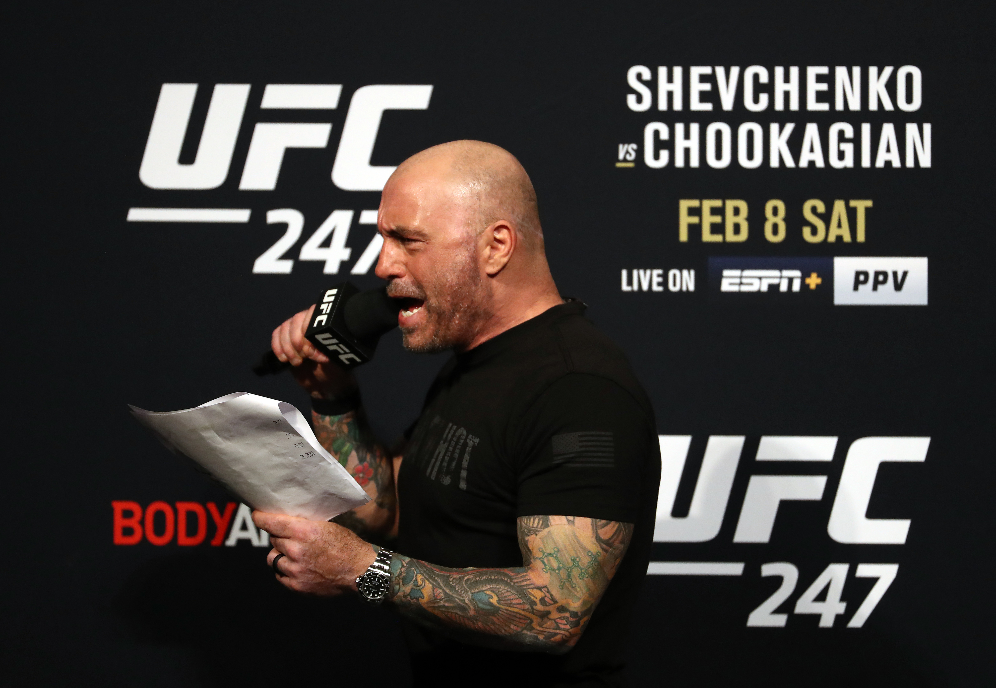 Joe Rogan speaks into a microphone while holding papers at the UFC 247 weigh-in event, with the fight card details in the background