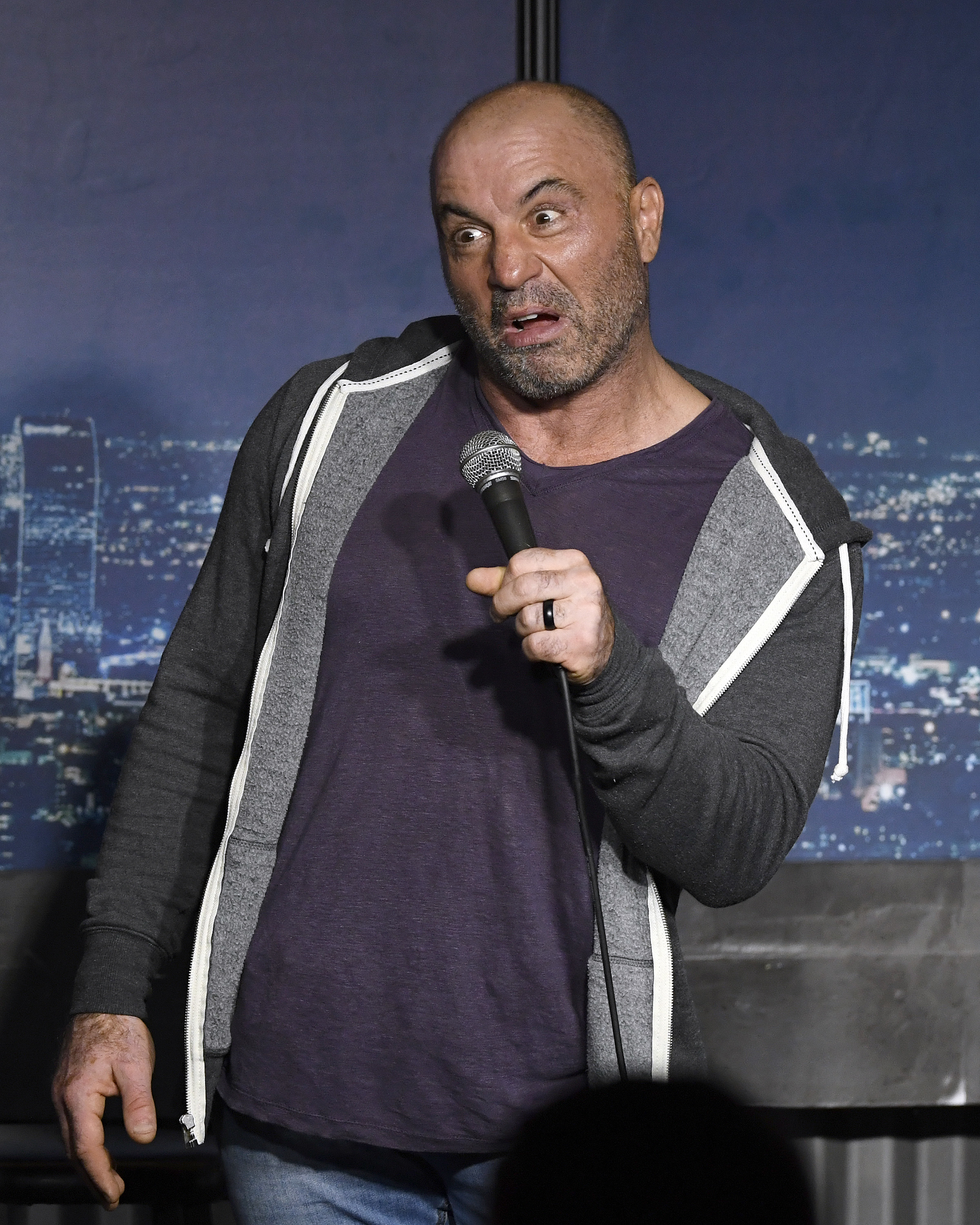 Joe Rogan, wearing a casual T-shirt and hoodie, holds a microphone and performs on stage with a cityscape background