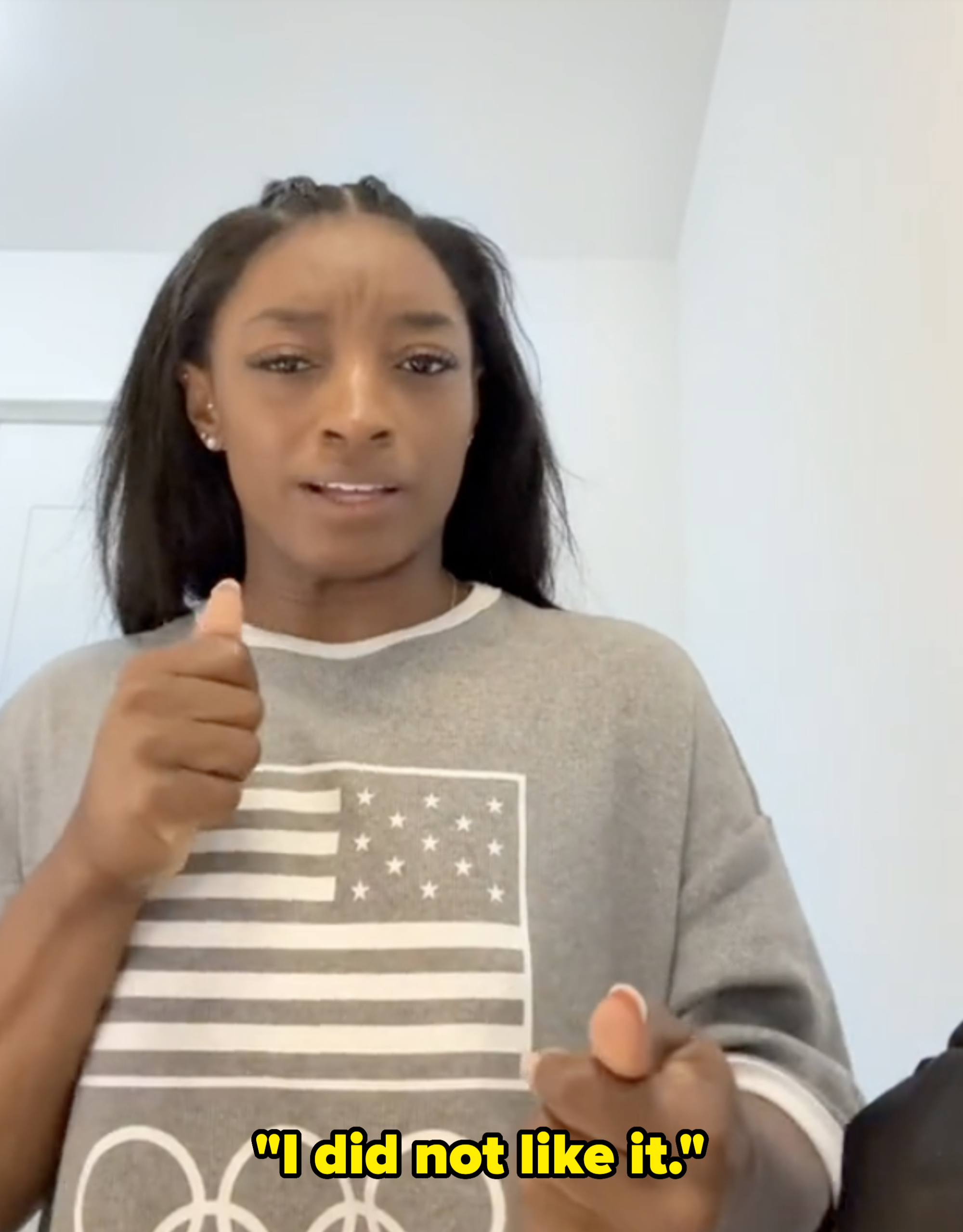 Simone Biles, wearing a casual shirt, makes a gesture with her right hand. The shirt features an American flag and Olympic rings
