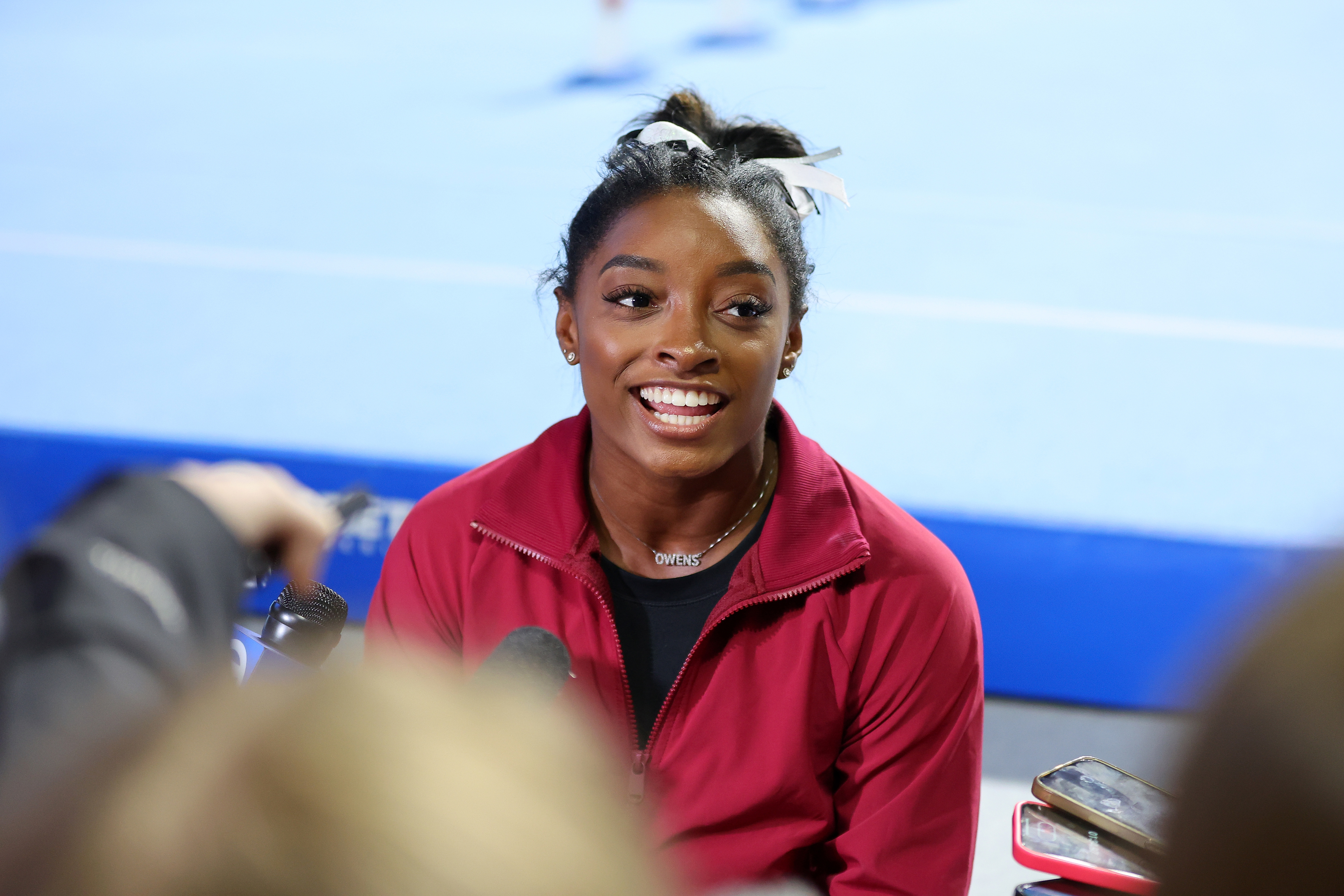 Simone Biles, wearing a red jacket, smiles while being interviewed by the press at a gymnastics event