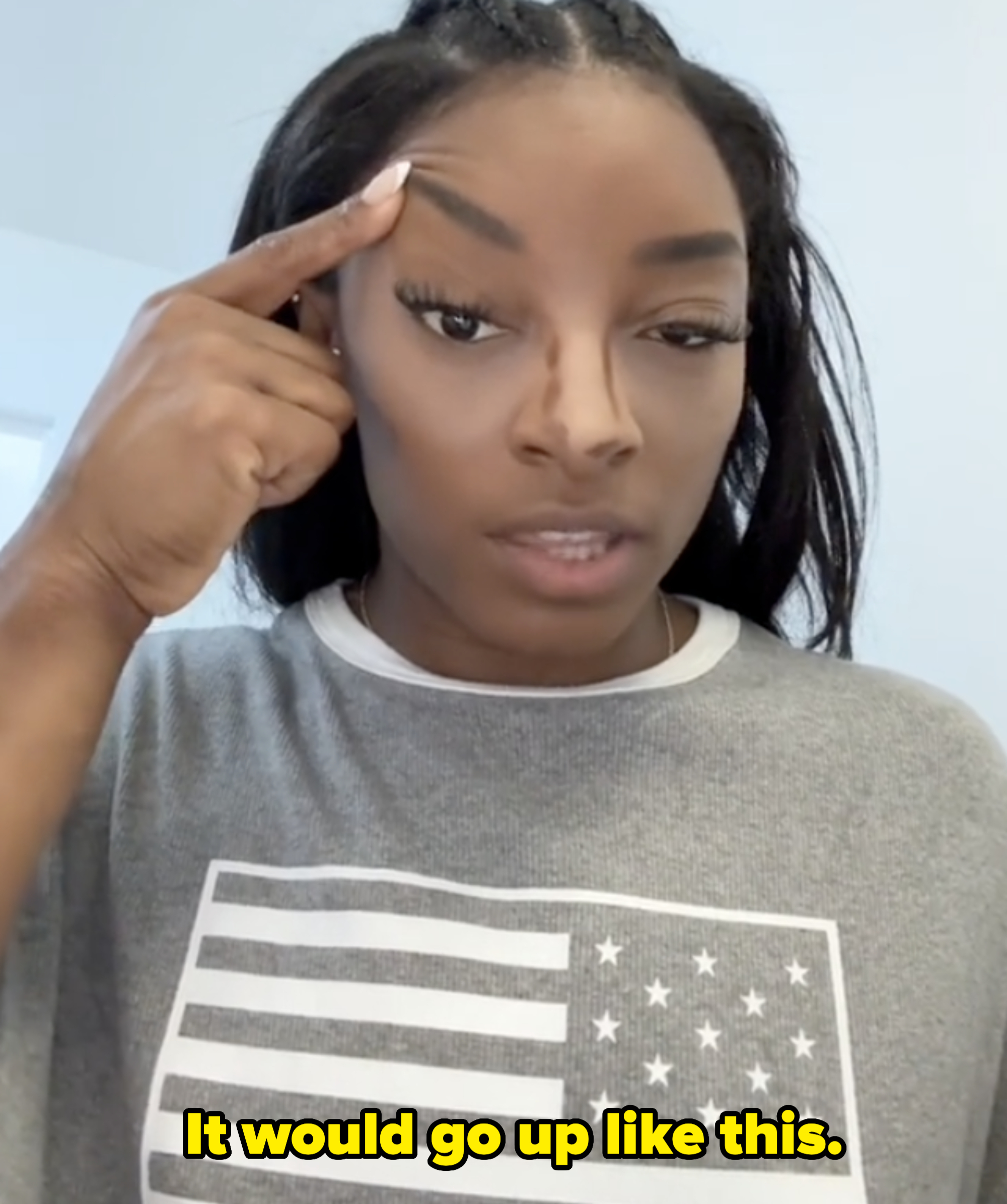 Simone Biles wearing a gray shirt with a flag design, pointing to her temple with her finger
