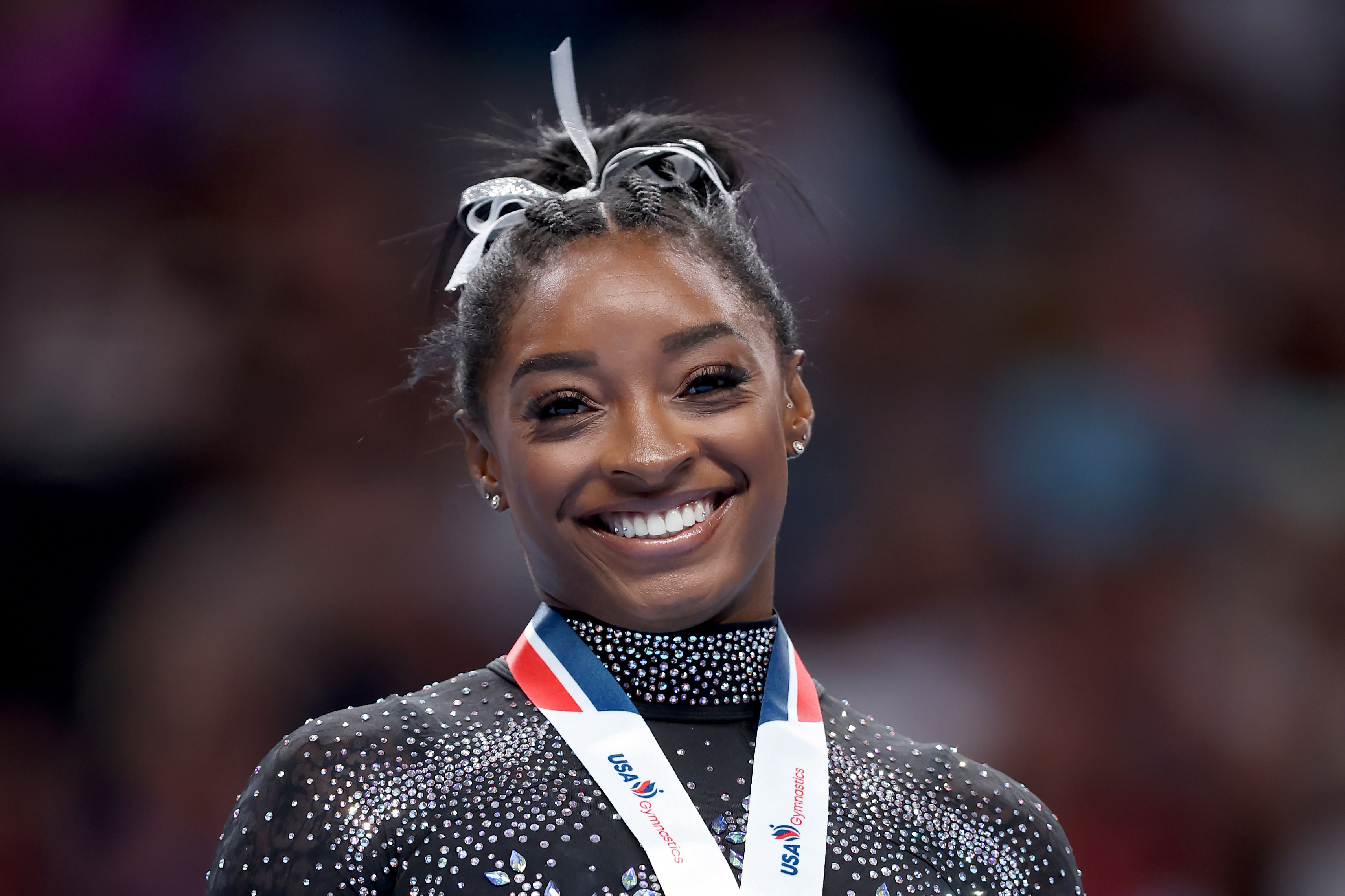 Simone Biles smiles at the camera, wearing a sparkly athletic outfit and a medal around her neck