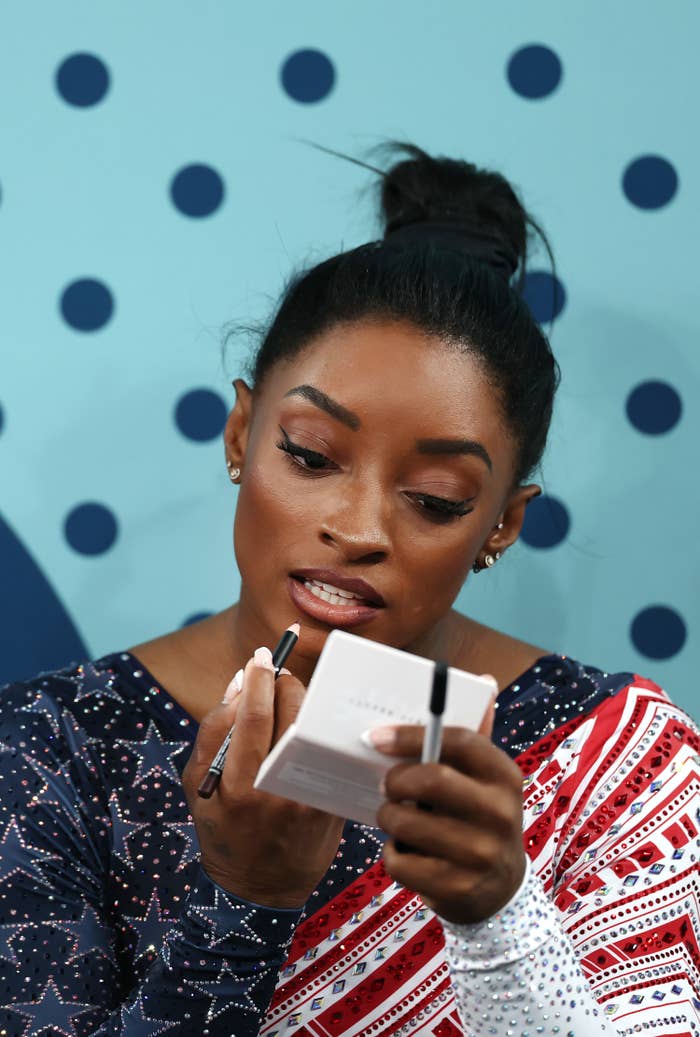 Simone Biles applies makeup while holding a compact mirror. She is wearing a stylish outfit featuring patterns and sequins