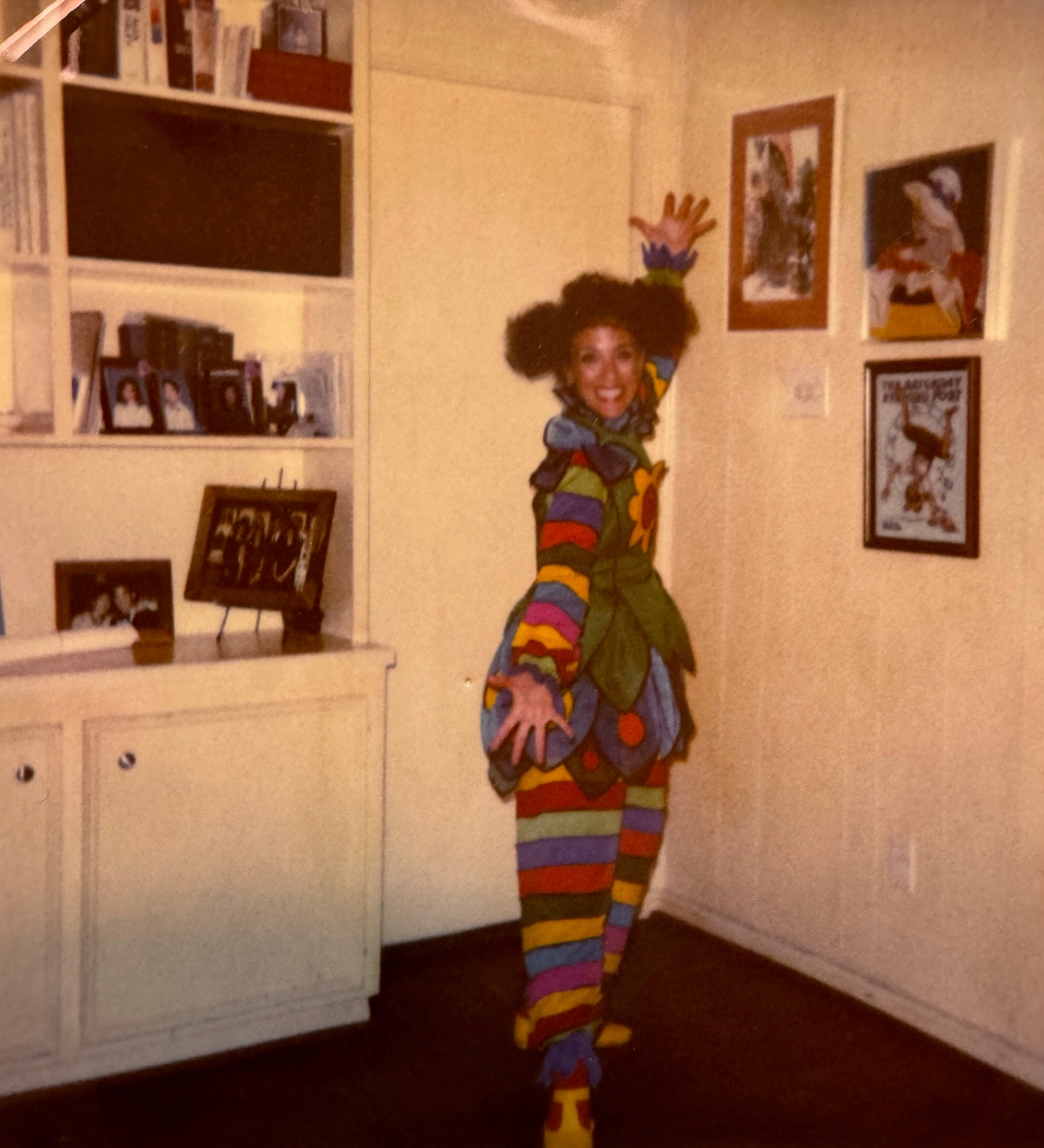Person in a colorful, striped costume with outstretched arms, standing indoors next to white cabinets and framed photos on the wall